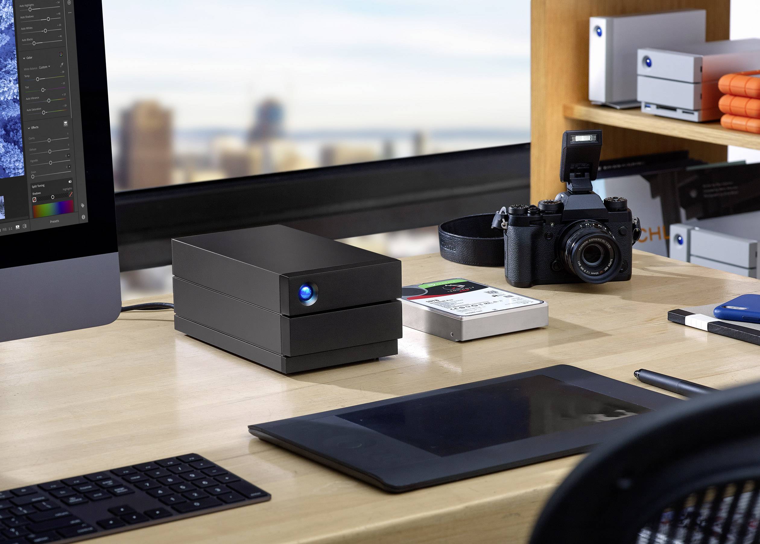 Een bureau met computermonitor, externe harde schijf, grafisch tablet en camera. Stadsaanzicht op de achtergrond door het raam.