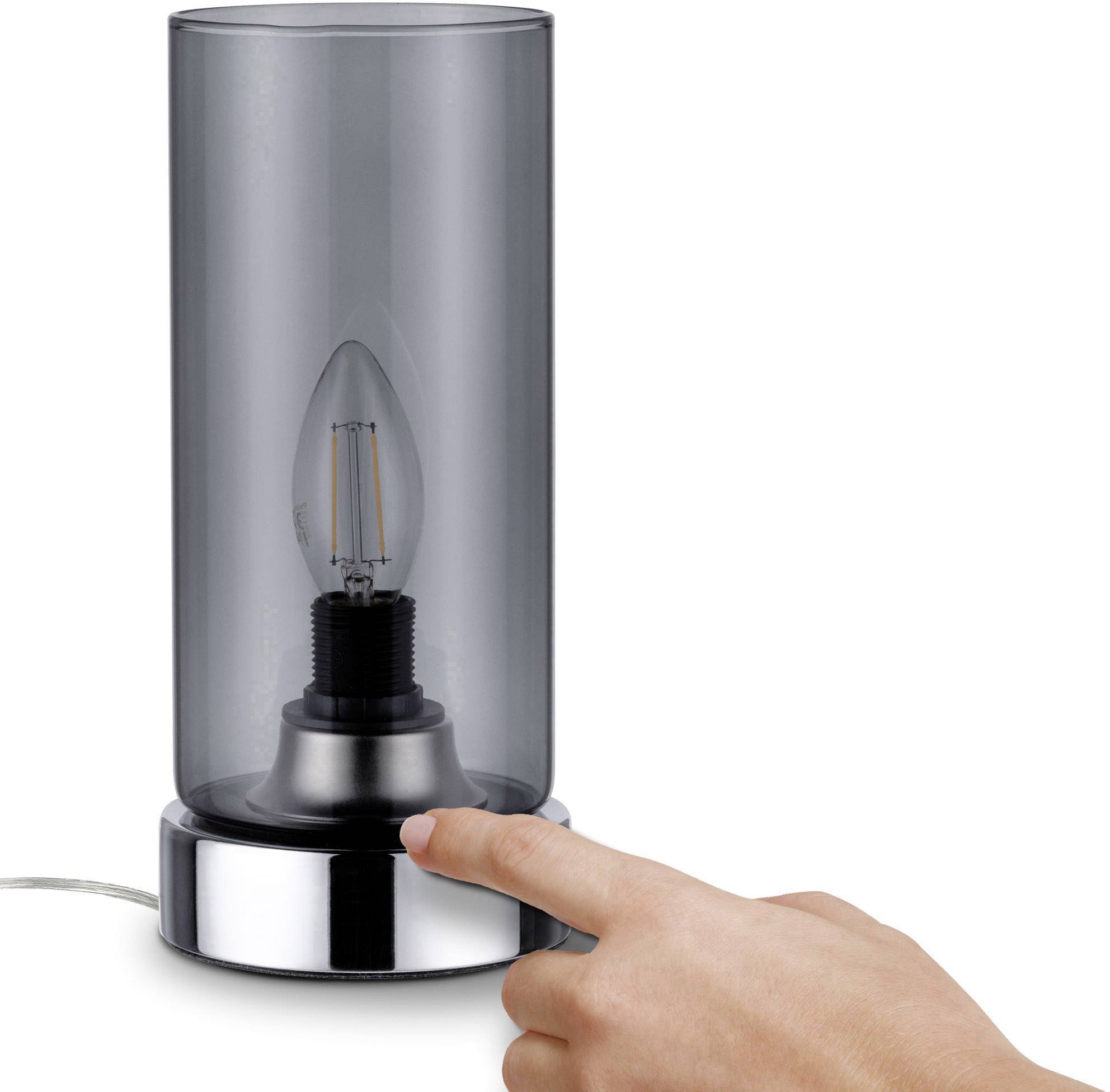 Glazen tafellamp met zichtbare gloeilamp in een cilindervorming lampenkap. Een hand raakt de voet van de lamp aan.