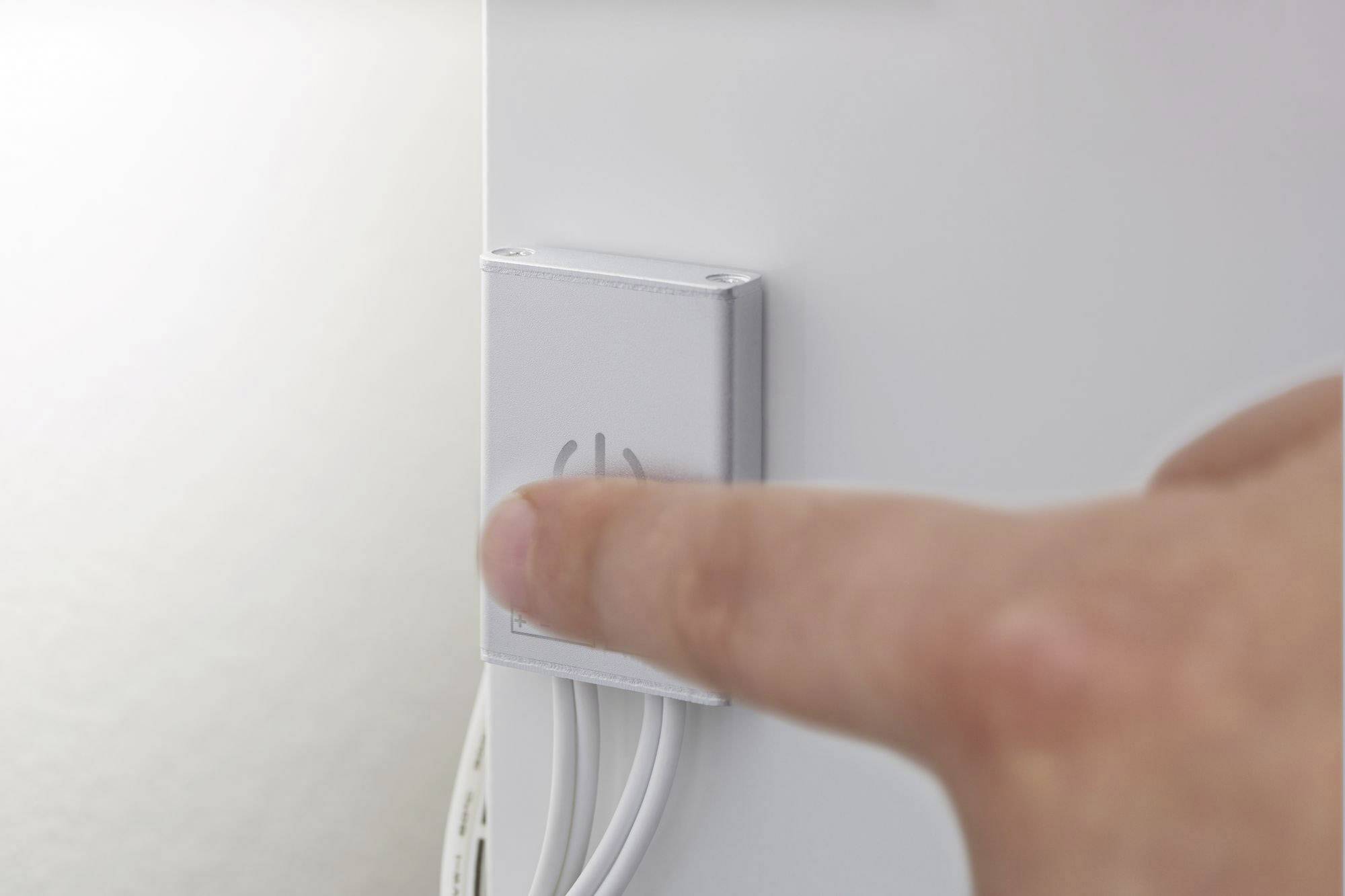 Een vinger drukt op een grote, zilveren knop aan de zijkant van een wit apparaat. Kabels zijn zichtbaar onder de knop.