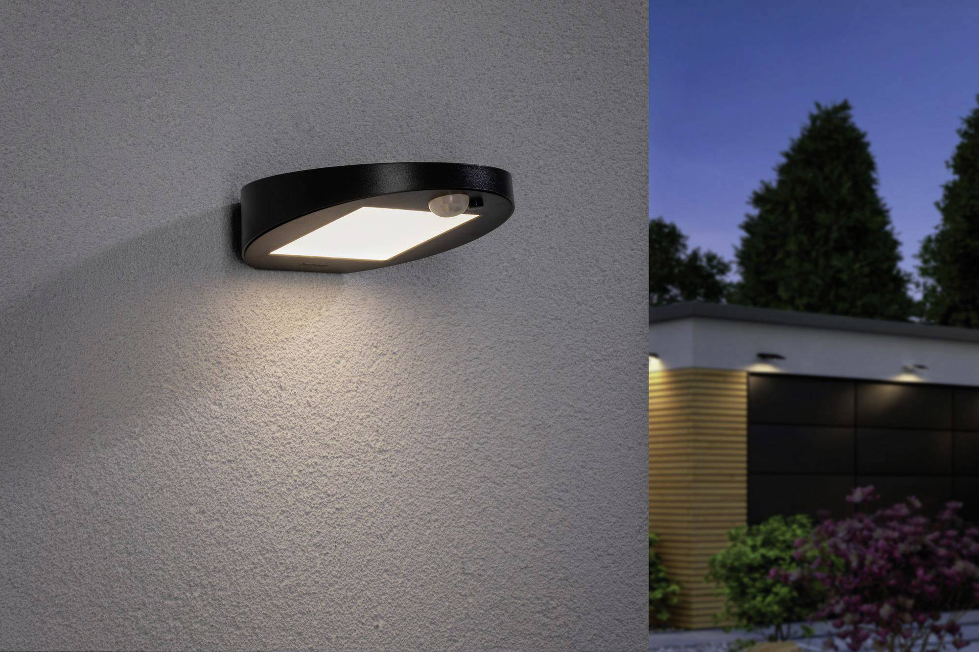 Moderne buitenlamp aan een grijze muur, werpt warm licht. Op de achtergrond een huis met bomen en een donkere lucht.