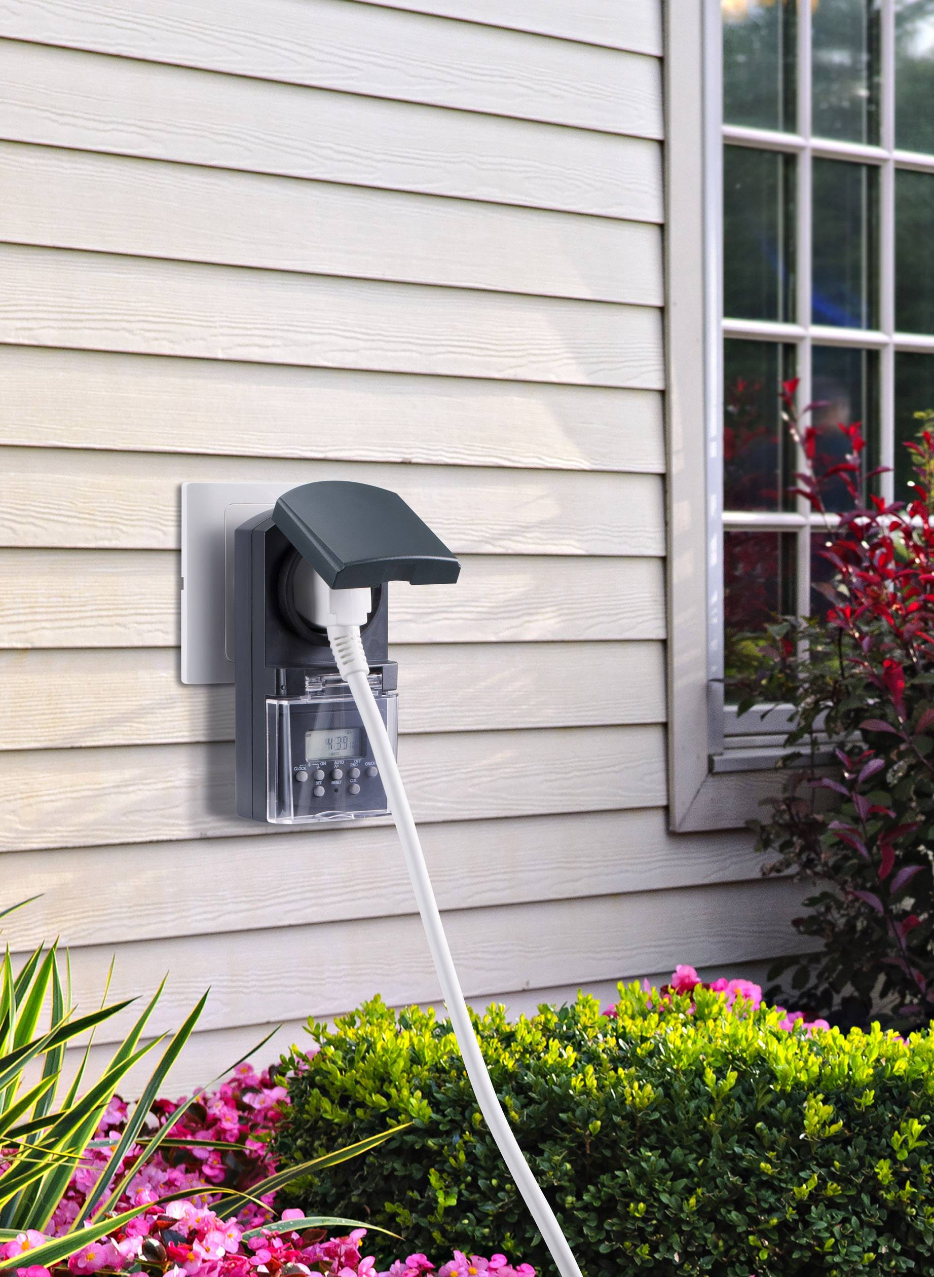 Buitenstopcontact aan een gevel met beschermkap. Een kabel is aangesloten, naast een raam en struiken.