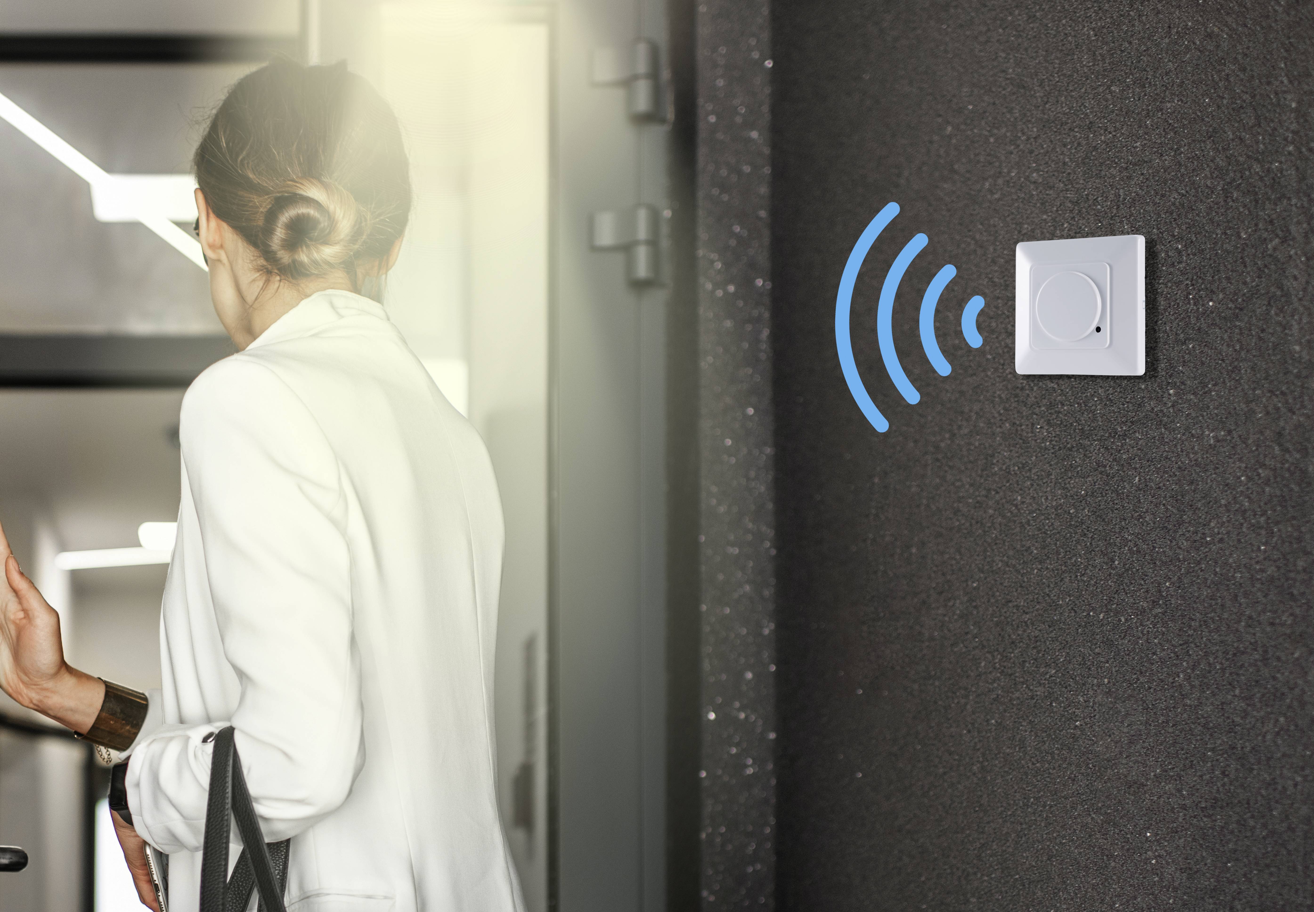 Een vrouw in een witte blazer betreedt een ruimte. Aan de muur is naast een lichtschakelaar een radiosignaal-symbool zichtbaar.