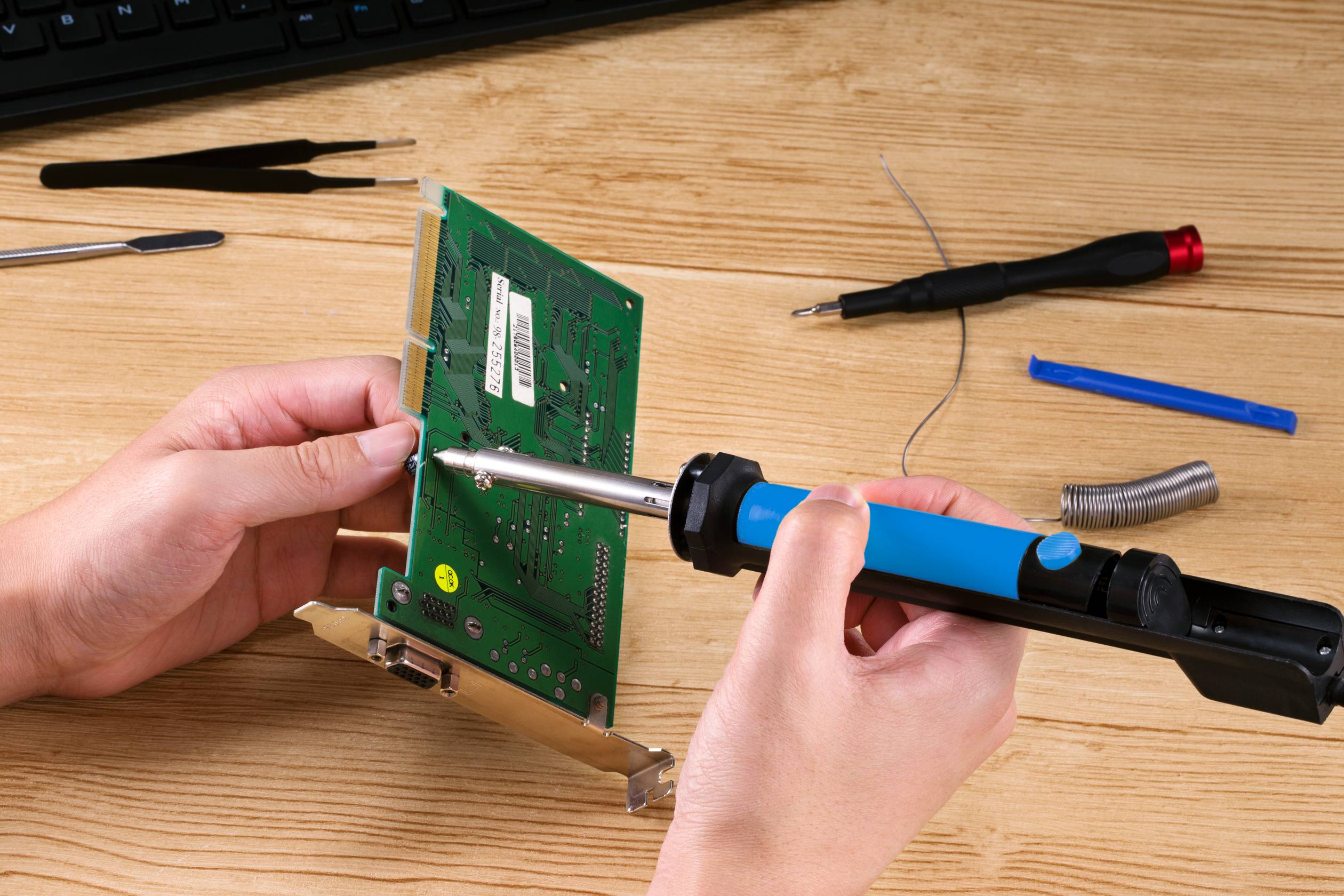 Een persoon soldeert een groene printplaat met een soldeerbout op een houten tafel, omringd door elektronicagereedschappen.