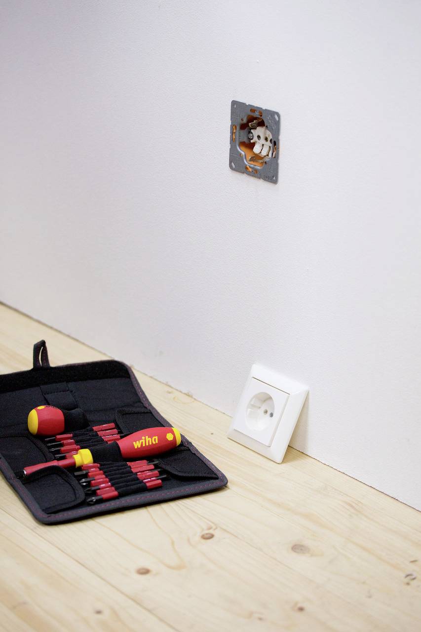 Een open stopcontact aan een muur, naast een gereedschapsset met schroevendraaiers op een houten vloer.