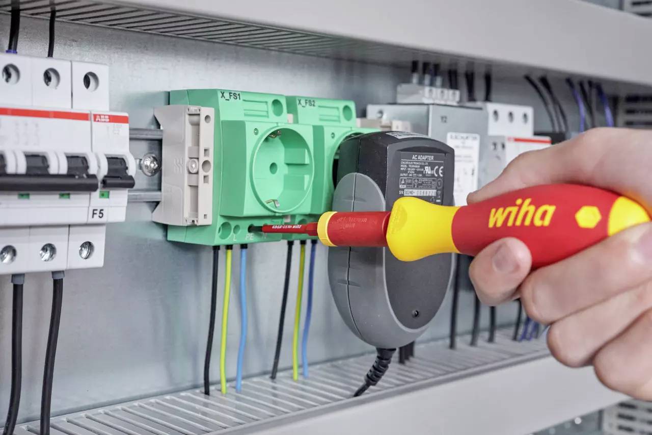 Een hand houdt een Wiha schroevendraaier vast terwijl draden in een elektrisch paneel worden beveiligd. Het paneel bevat verschillende schakelaars en circuitcomponenten.
