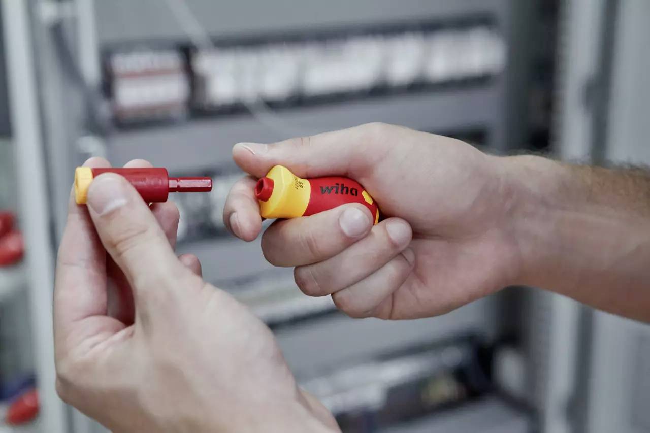 Twee handen die een rood en geel gereedschap met een afneembare tip, mogelijk een schroevendraaier, vasthouden voor een wazig elektrisch paneel.