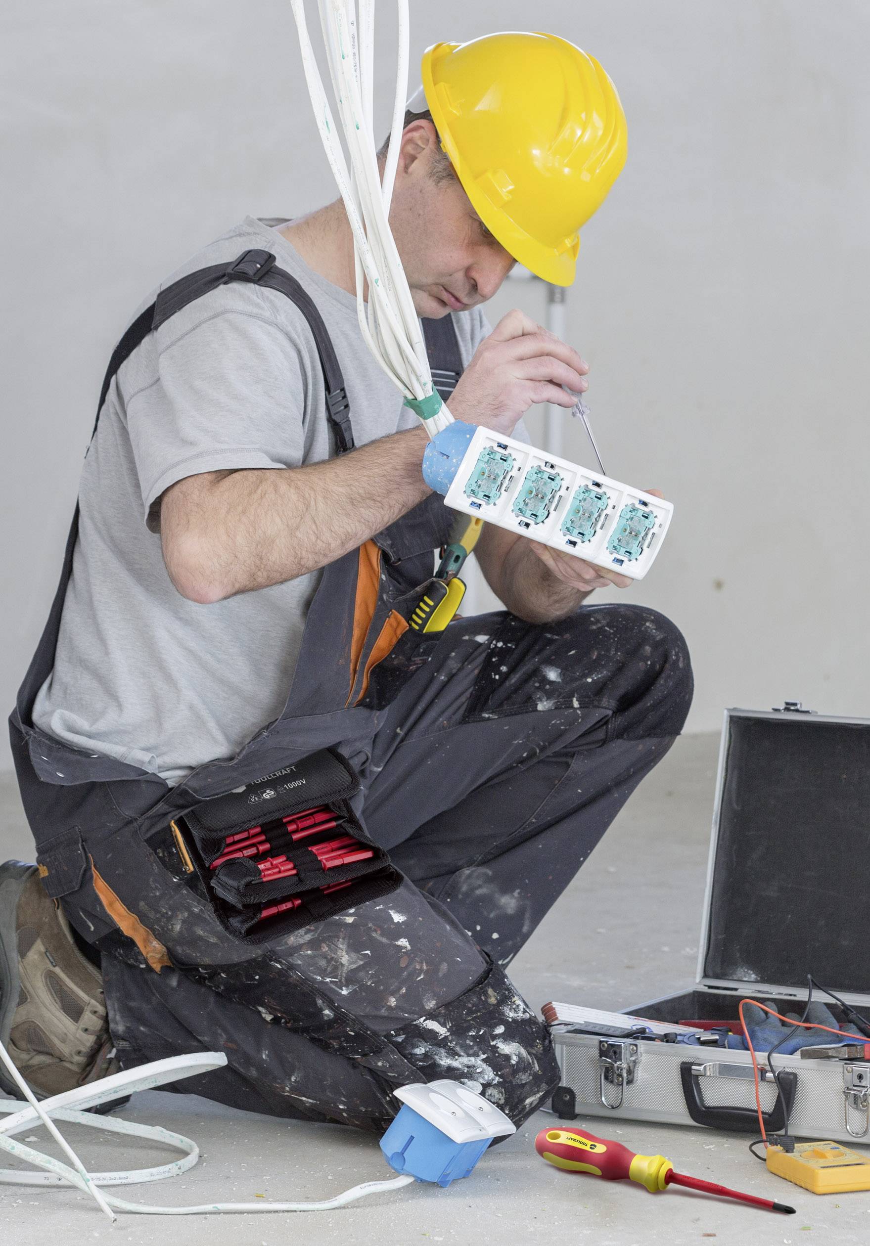 Een elektricien met een gele helm werkt aan een schakelkast, omringd door gereedschappen. Hij concentreert zich op de bedrading.