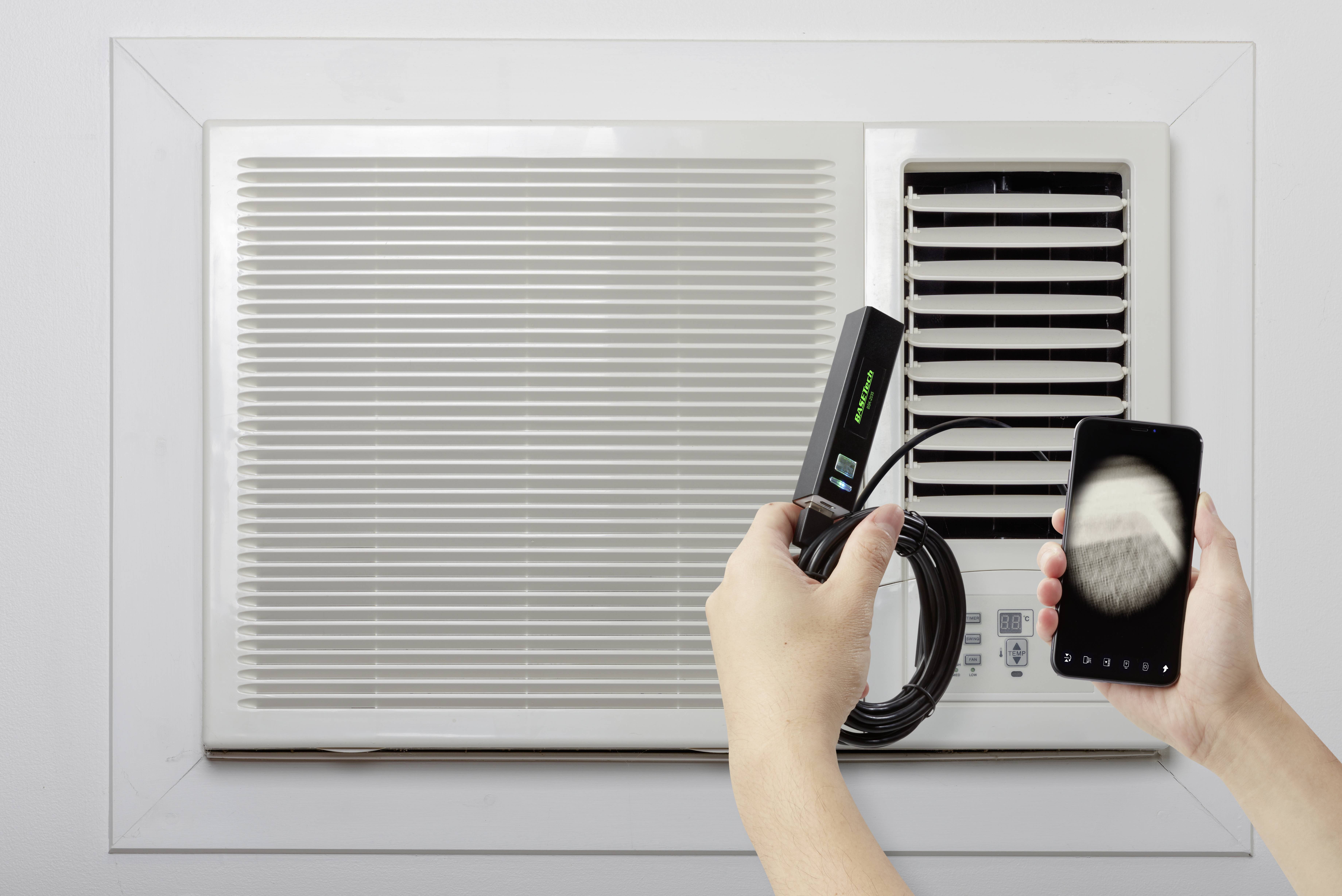 Een persoon houdt een smartphone en een bekabeld apparaat dicht bij een airconditioning. De handeling wijst op een verbinding of meting.