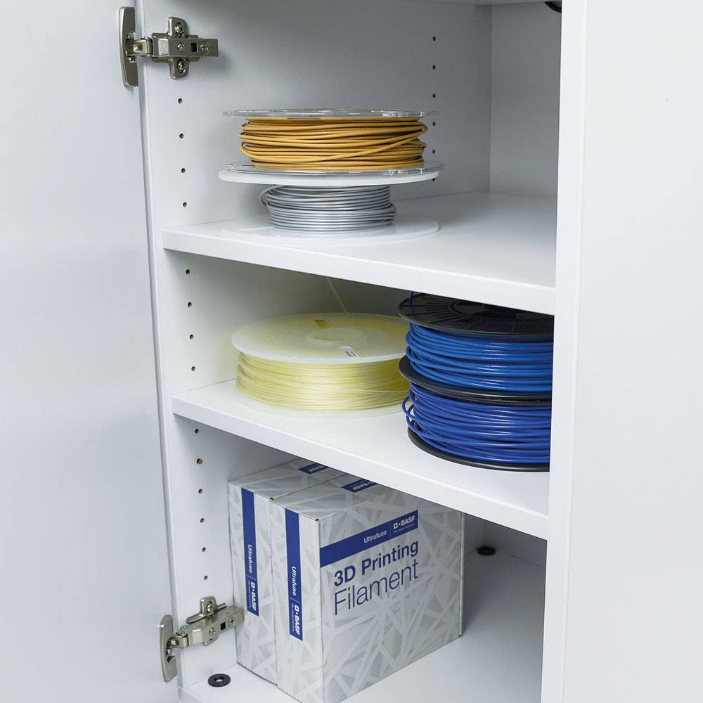Een open kast toont verschillende rollen 3D-printer-filamenten in de kleuren oranje, geel, wit en blauw, samen met een doos.