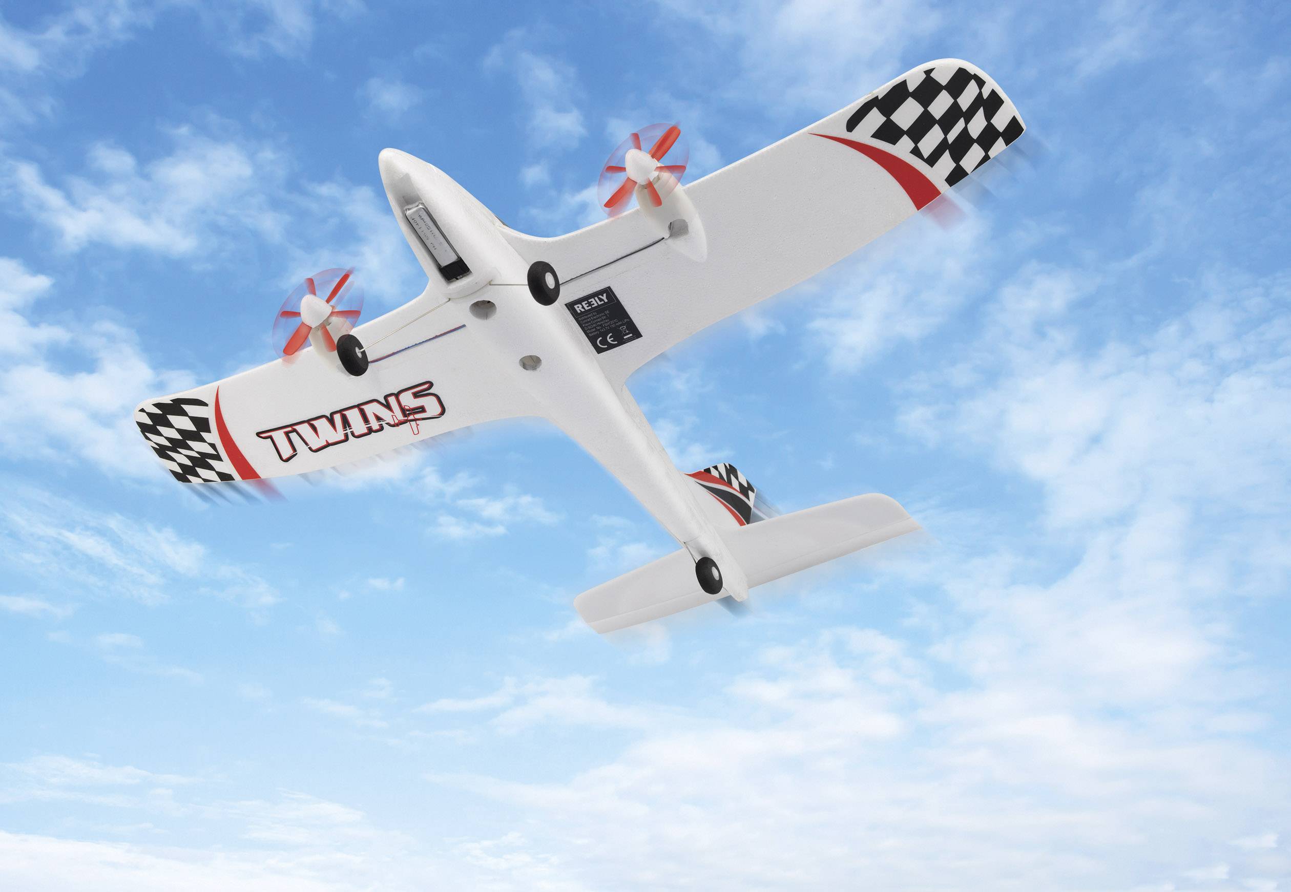 Speelgoedvliegtuig met zwart-wit geruit patroon en de tekst 'TWINS' vliegt in een heldere blauwe lucht.