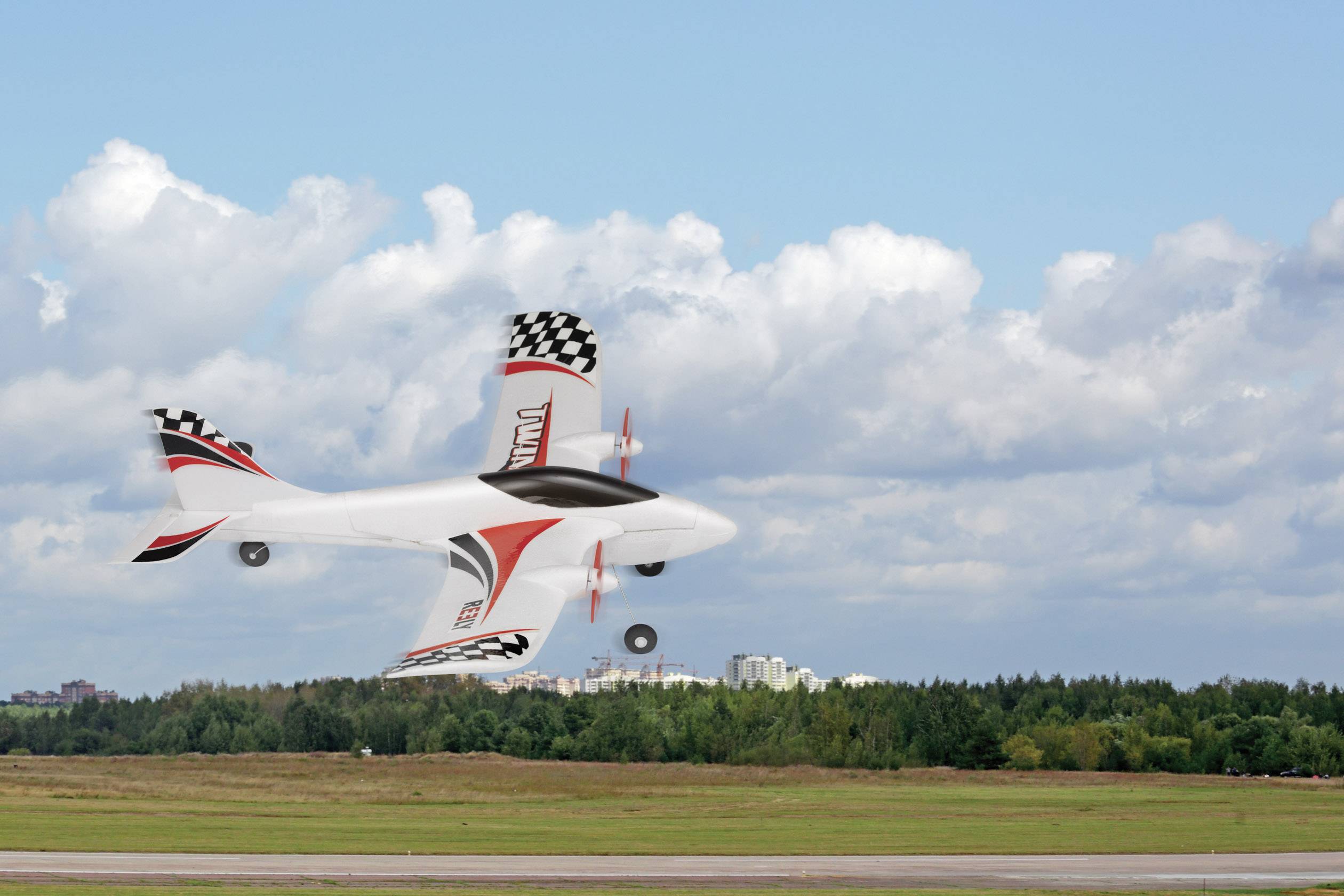 Een op afstand bestuurbaar modelvliegtuig in rood en wit zweeft in de lucht boven een groene weide met bomen en een bewolkte lucht.