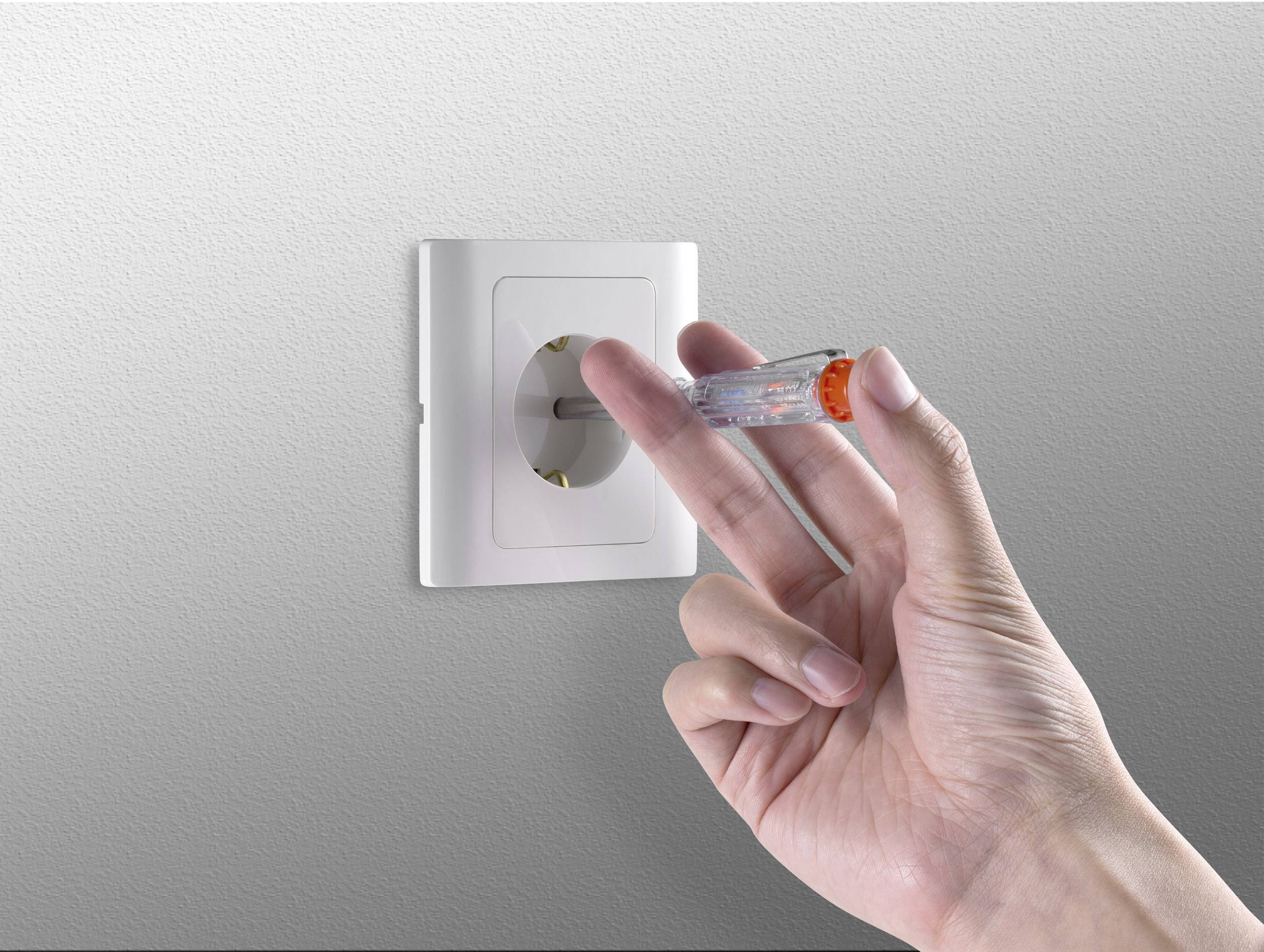 Een hand houdt een spanningszoeker dicht bij een witte stopcontact aan een grijze muur om elektrische spanning te controleren.