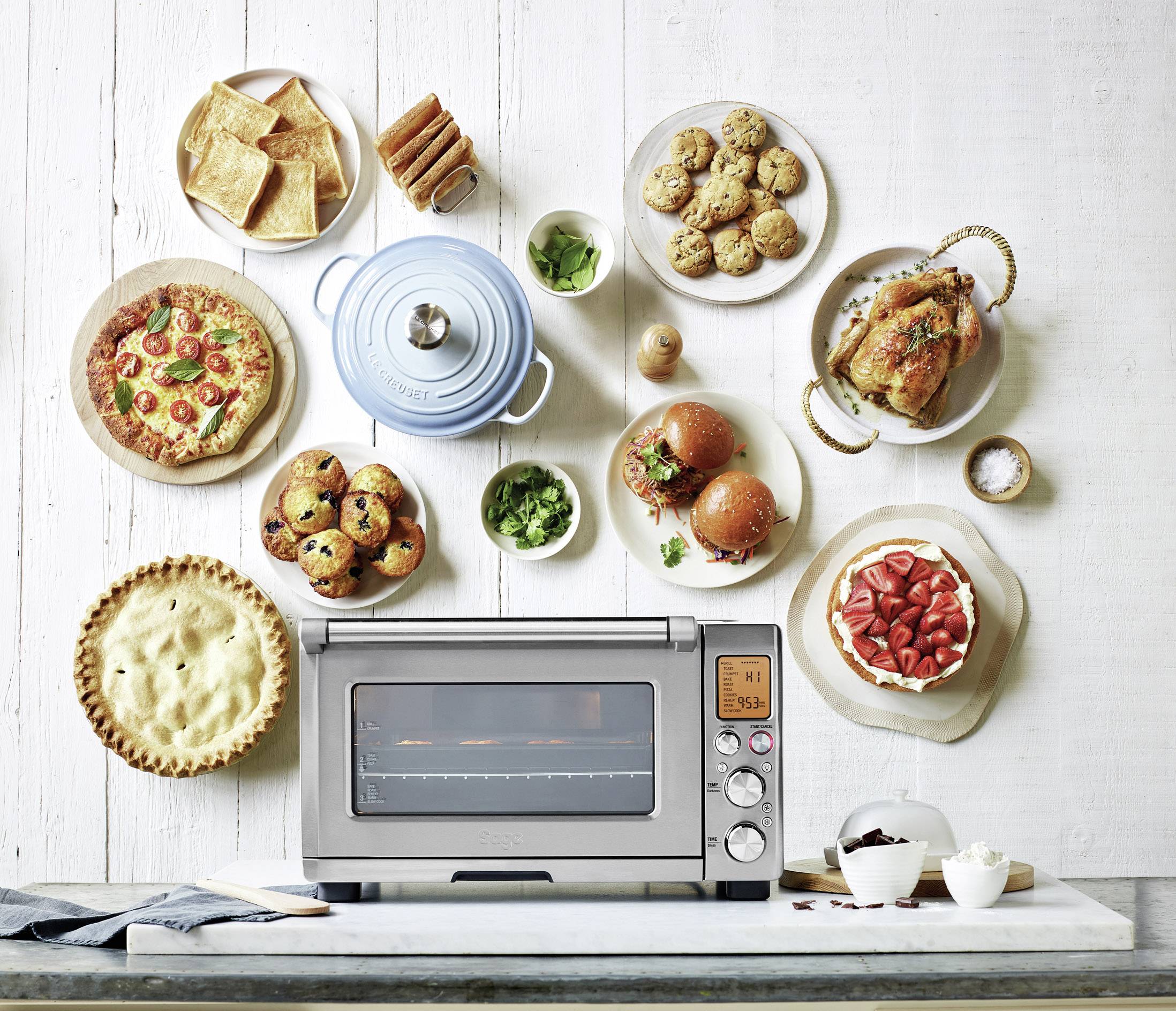 Multifunctionele oven op een tafel, omringd door gebak, pizza, geroosterde kip en salade. Toont de kookmogelijkheden van de oven.