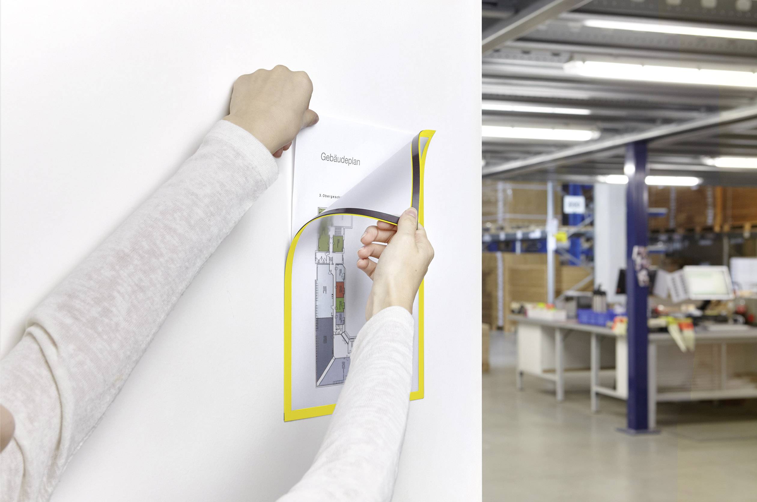 Een persoon hangt een poster aan een bureaumuur. Op de achtergrond is een werkruimte met bureaus en planken te zien.