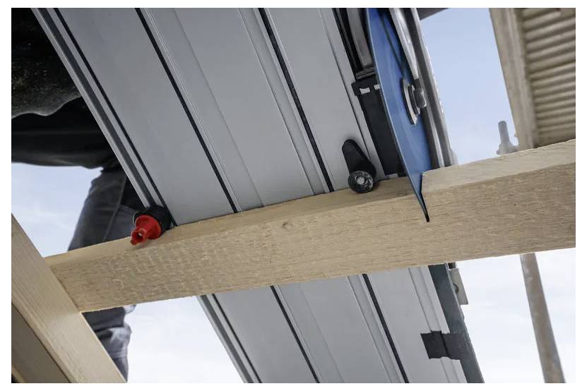 Een close-up van een timmerman die hout zaagt met een geleiderailzaag, met de nadruk op de geleiderail en de houten plank. De lucht is zichtbaar op de achtergrond.