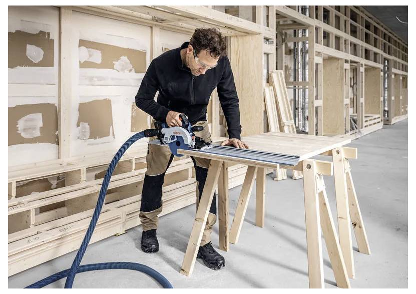 Een persoon in een werkplaats gebruikt een elektrisch gereedschap om een houten plank door te zagen op een schraag, omringd door onafgewerkte houten kozijnen en gipsplaten.