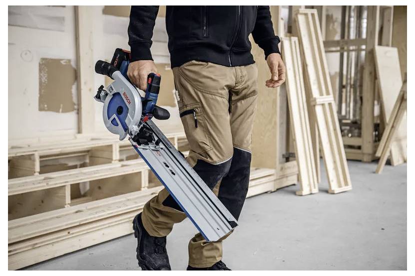 Een persoon in een werkplaats houdt een snoergeleide elektrische cirkelzaag vast, terwijl hij langs houten planken loopt. Ze dragen werkbroek en zwarte laarzen, wat wijst op timmerwerk.