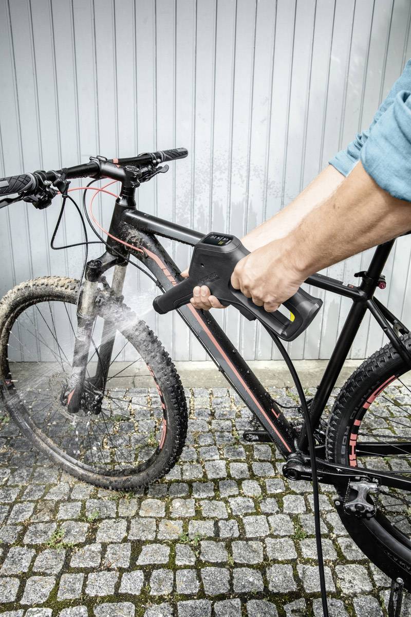 Een persoon reinigt een modderig mountainbike met een hogedrukreiniger op een bestrate ondergrond voor een grijze muur.