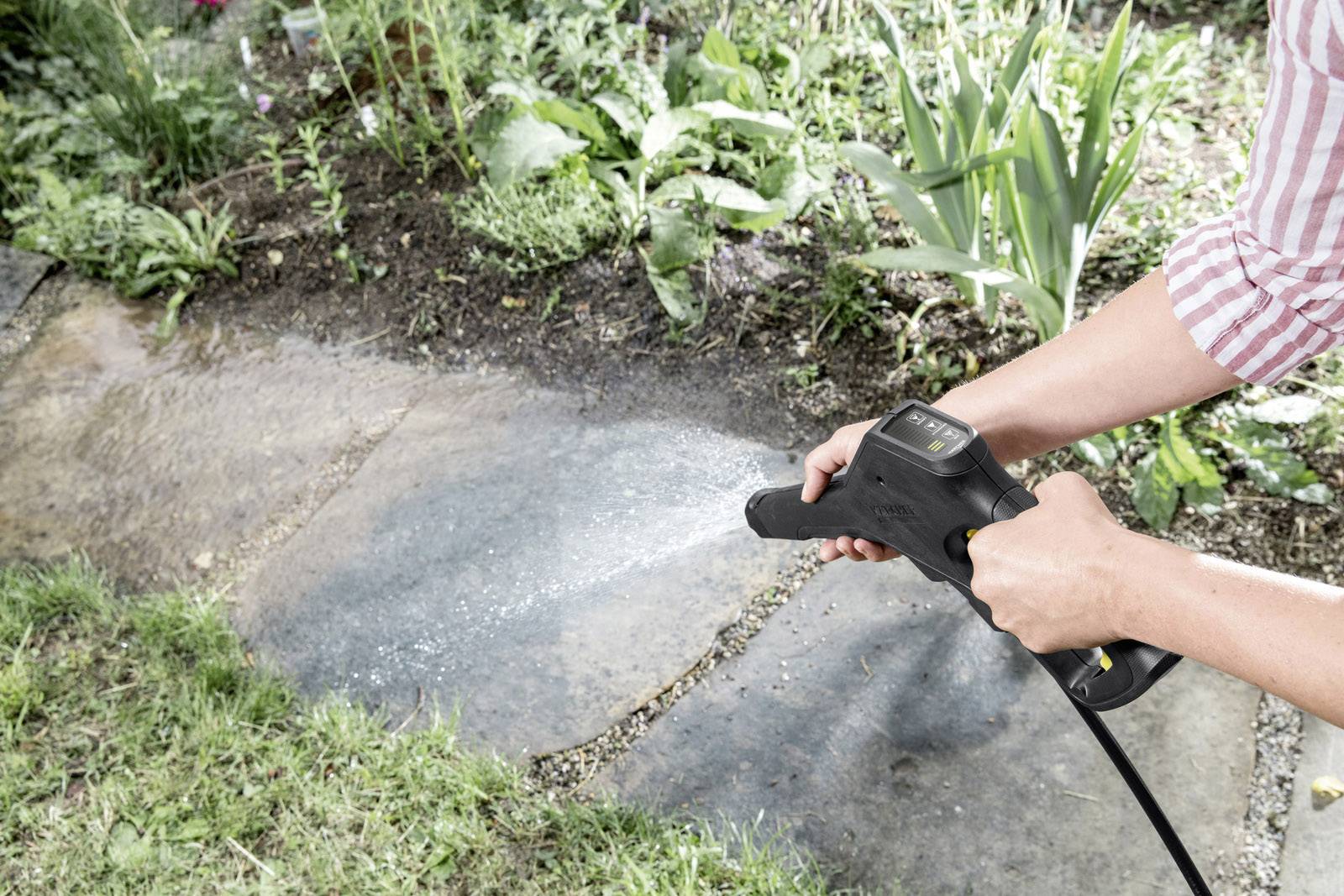 Een persoon reinigt een terrastegel met een hogedrukreiniger. Omgeven door gras en planten.