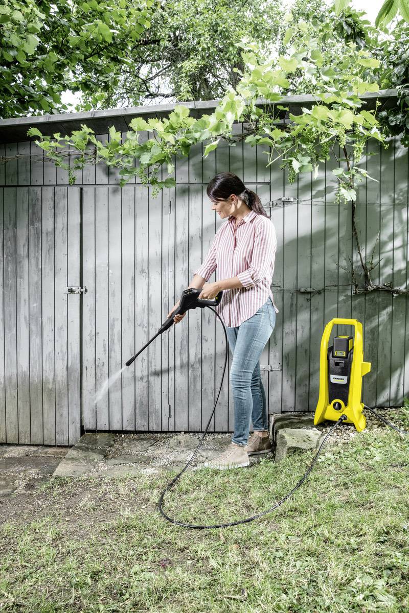 Een persoon reinigt de muren van een schuur in de tuin met een hogedrukreiniger. Op de achtergrond zijn bomen zichtbaar.