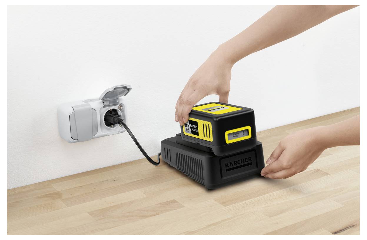 Handen die een zwarte en gele batterij op een oplader plaatsen op een houten oppervlak, waarbij de oplader is aangesloten op een stopcontact.