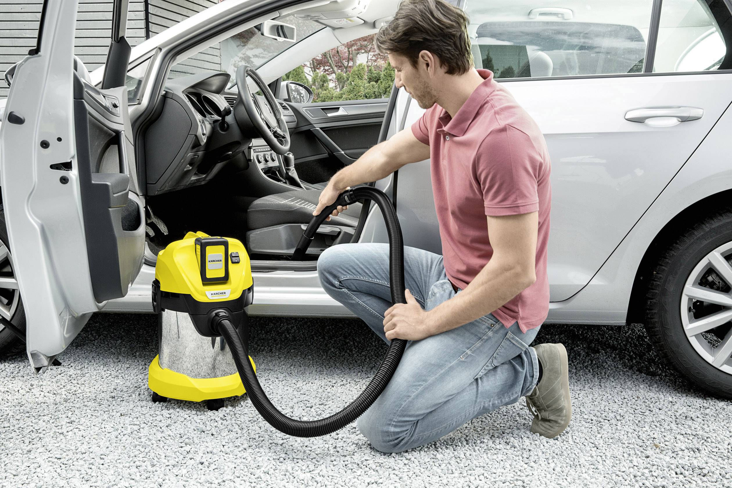 Een man knielt naast een zilveren auto en reinigt het interieur met een gele stofzuiger.