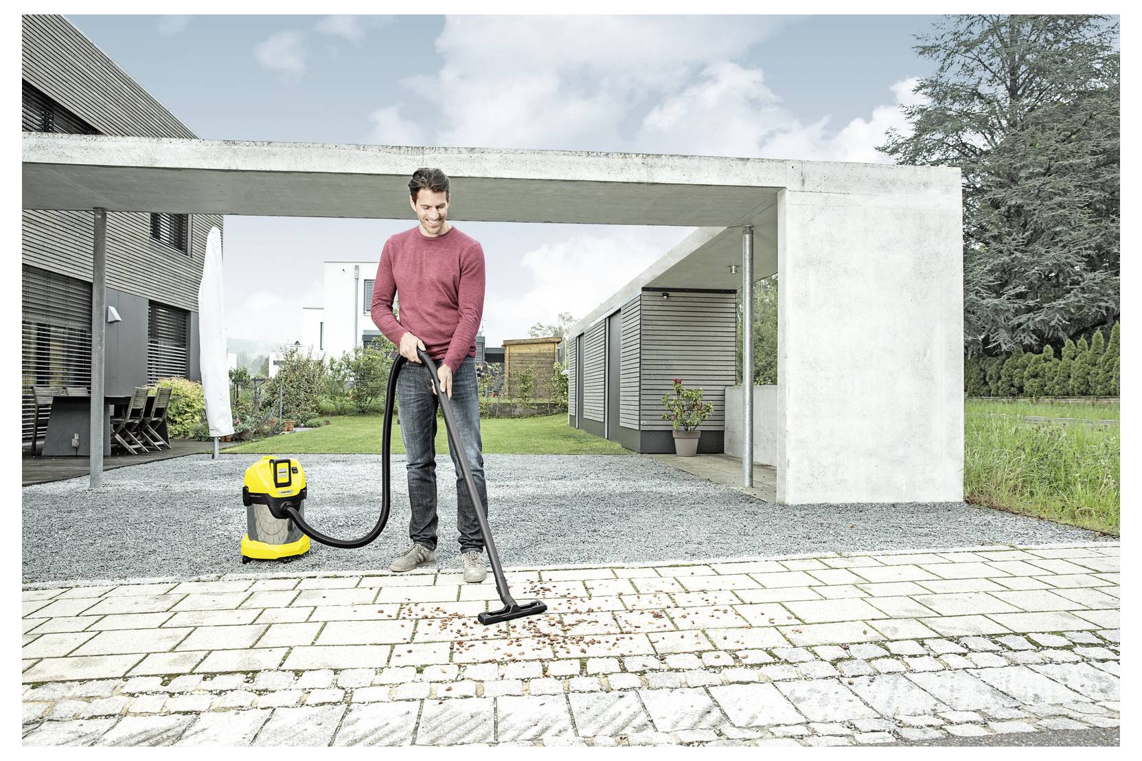 Een persoon zuigt gevallen bladeren van een bestrate patio met een geel en zwarte stofzuiger op de oprit van een modern huis.