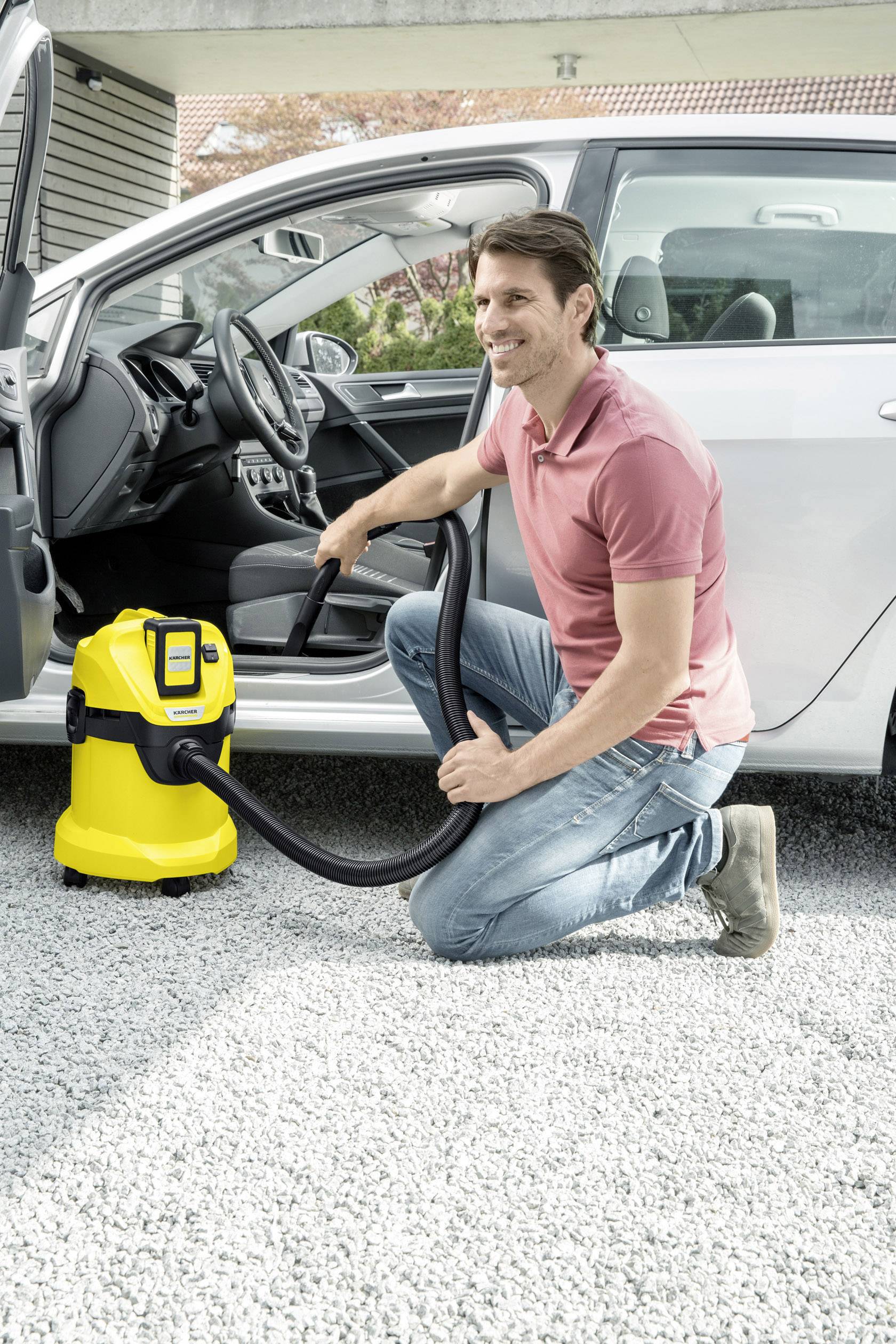 Een man knielt naast een zilveren auto en gebruikt een gele industriële stofzuiger om het interieur schoon te maken.