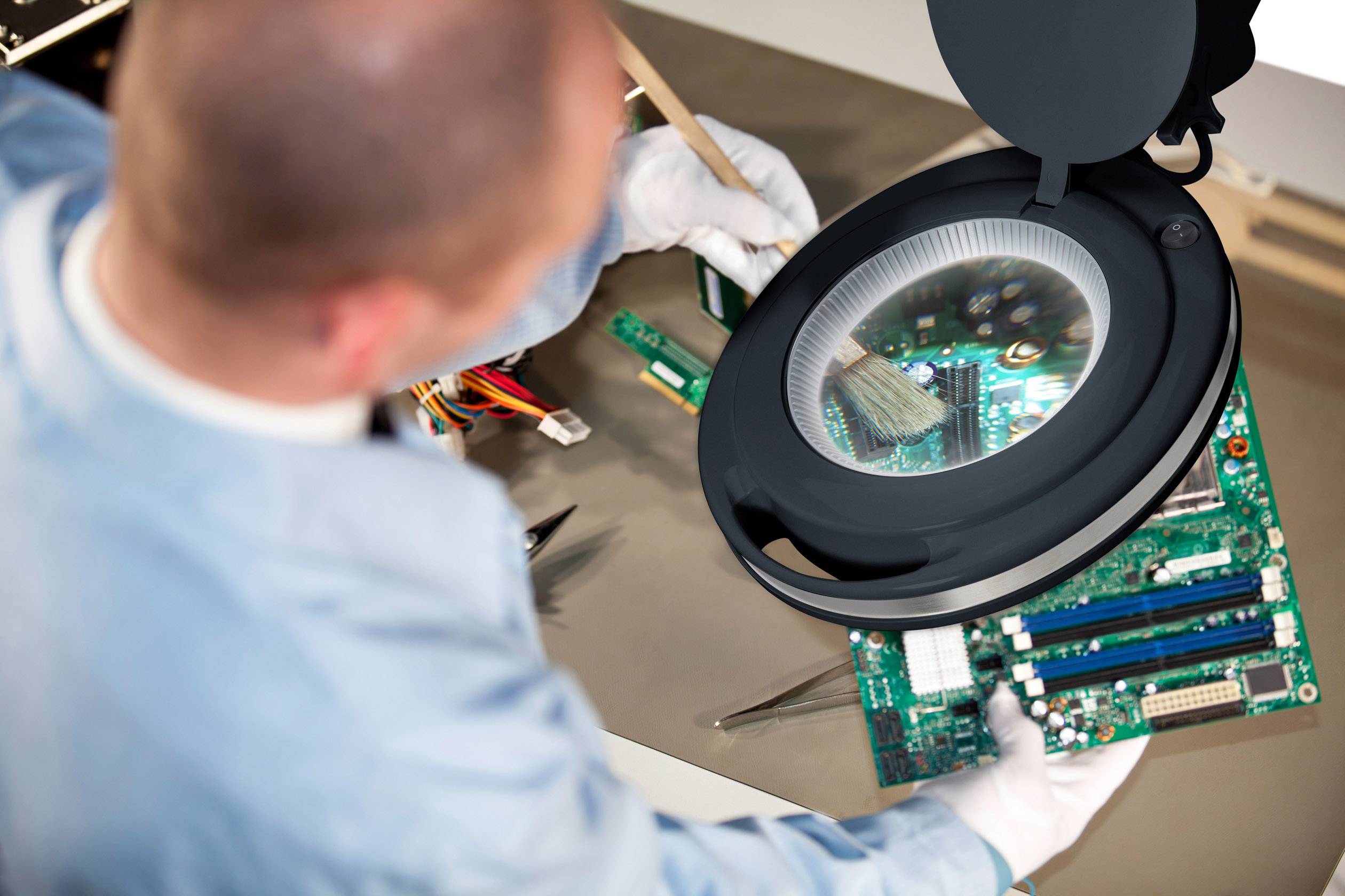 Een technicus reinigt een open computerprint met een penseel onder een verlichte loep op een tafel.