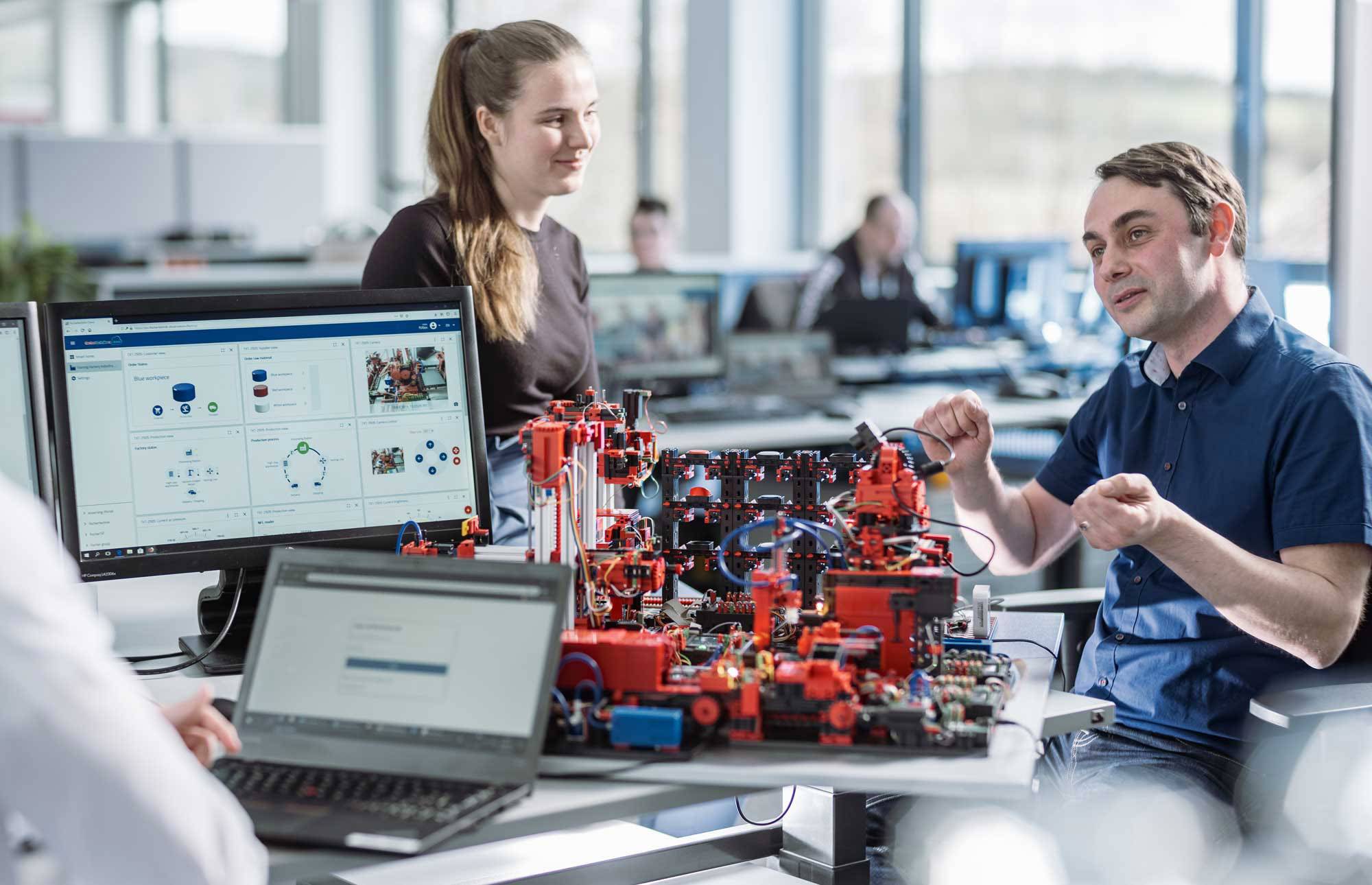 Drie personen discussiëren over een technisch model op een tafel in een kantoor. Op de monitors zijn diagrammen en gegevens te zien.