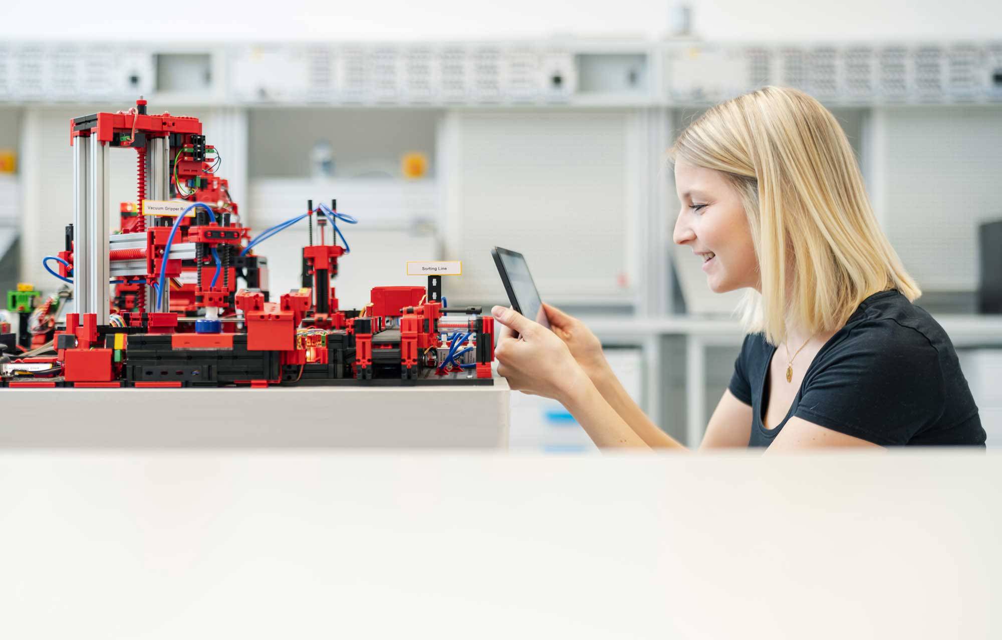 Een vrouw werkt geconcentreerd aan een tafel met een smartphone, naast haar staat een complex model van rode bouwstenen in een moderne ruimte.