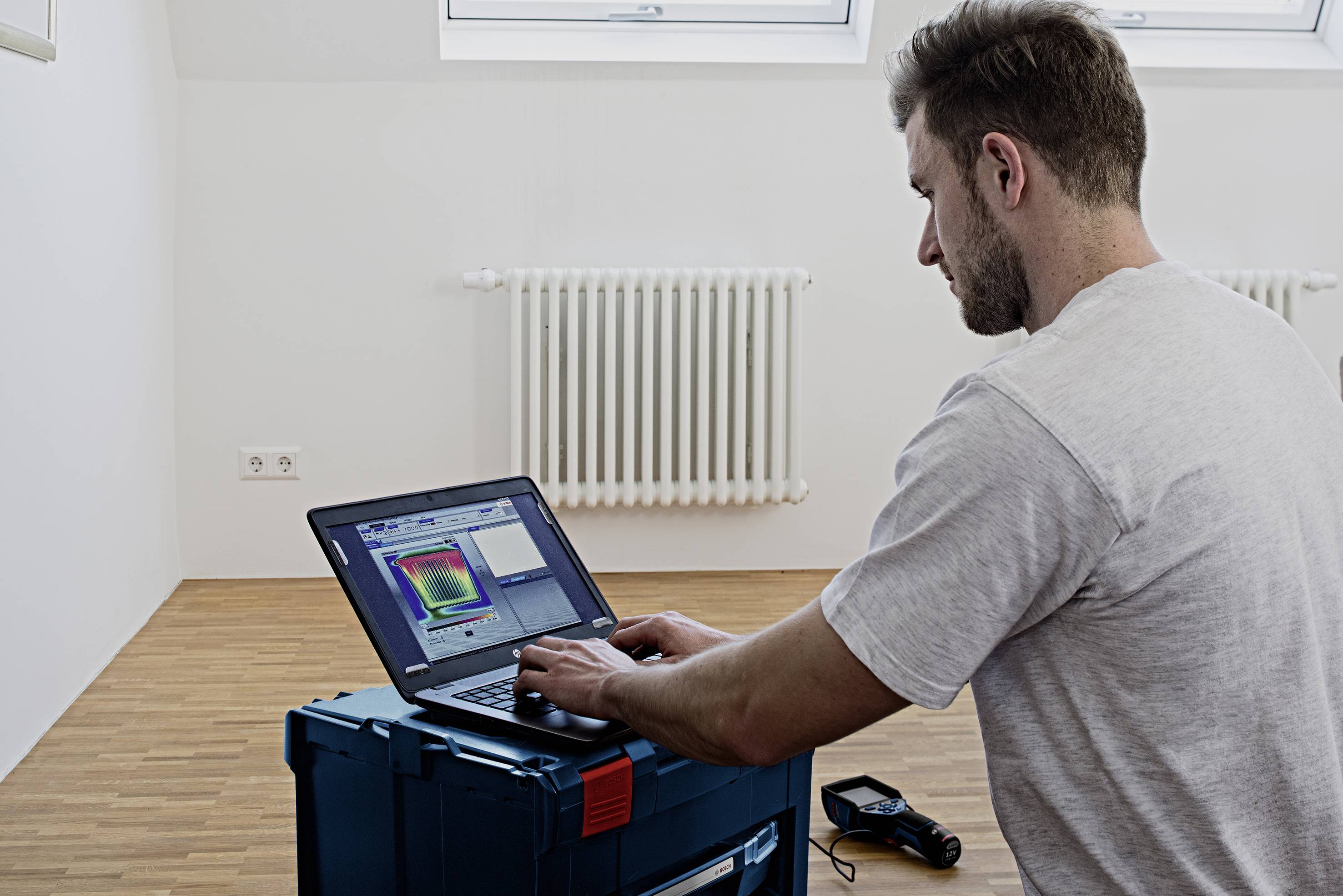 Een man werkt aan een laptop die op een gereedschapskist staat. Het scherm toont gekleurde diagrammen. Op de achtergrond is een radiator zichtbaar.