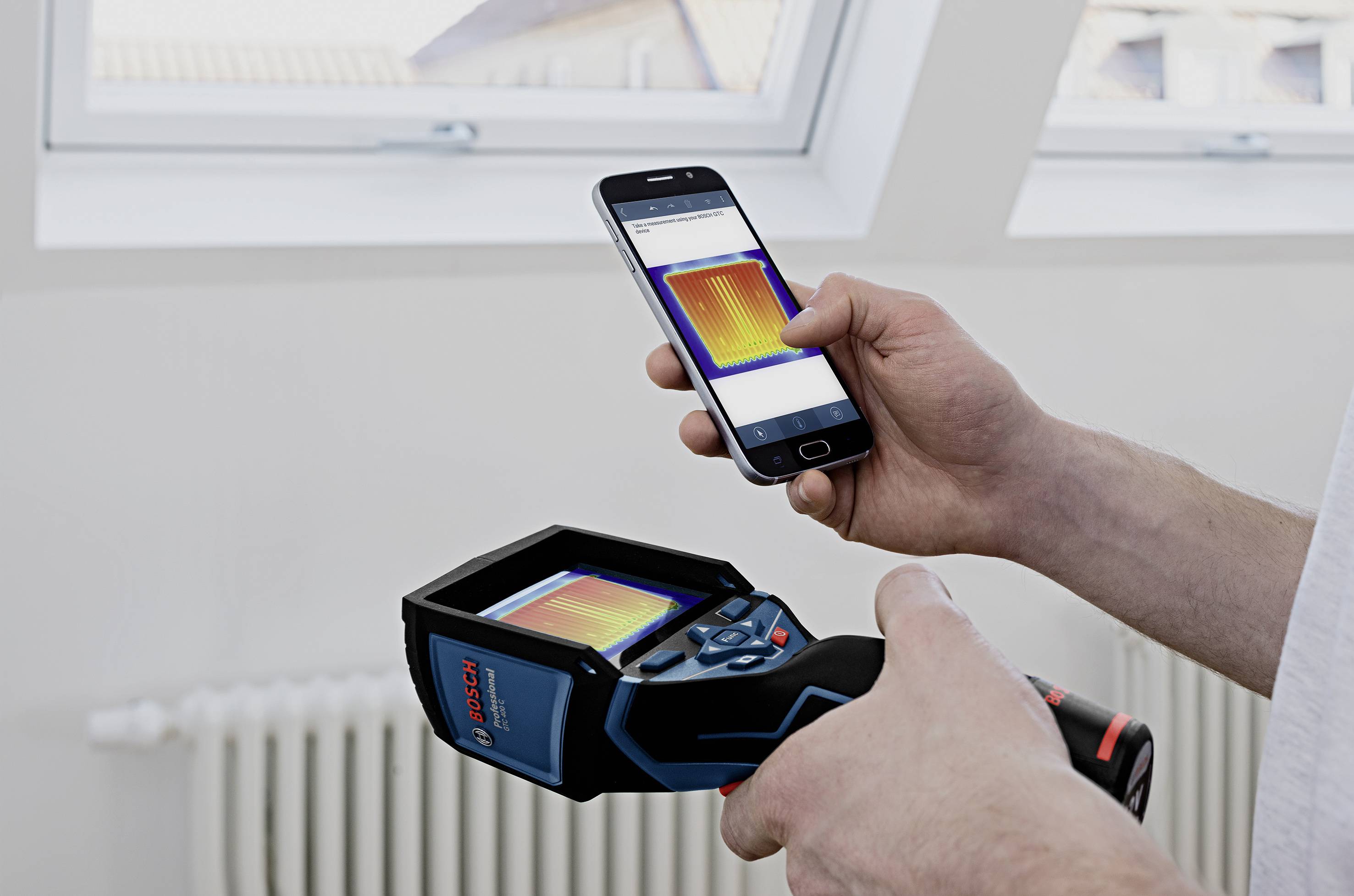 Een persoon houdt een warmtebeeldcamera en een smartphone vast. Beide displays tonen een warmtebeeld. Op de achtergrond is een radiator zichtbaar.