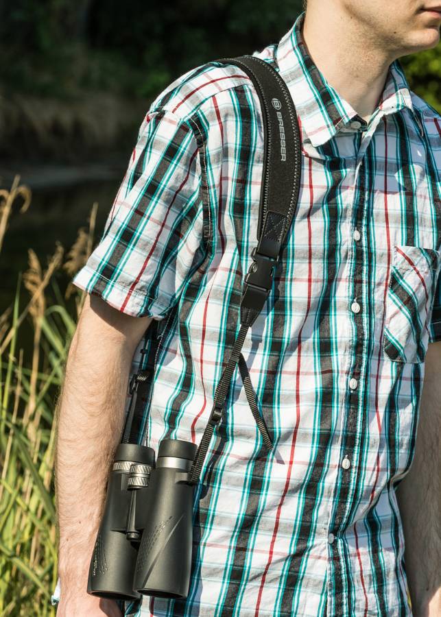 Een persoon in een geruit overhemd draagt een verrekijker om de schouder, staand in een natuurlijke omgeving met hoog gras.