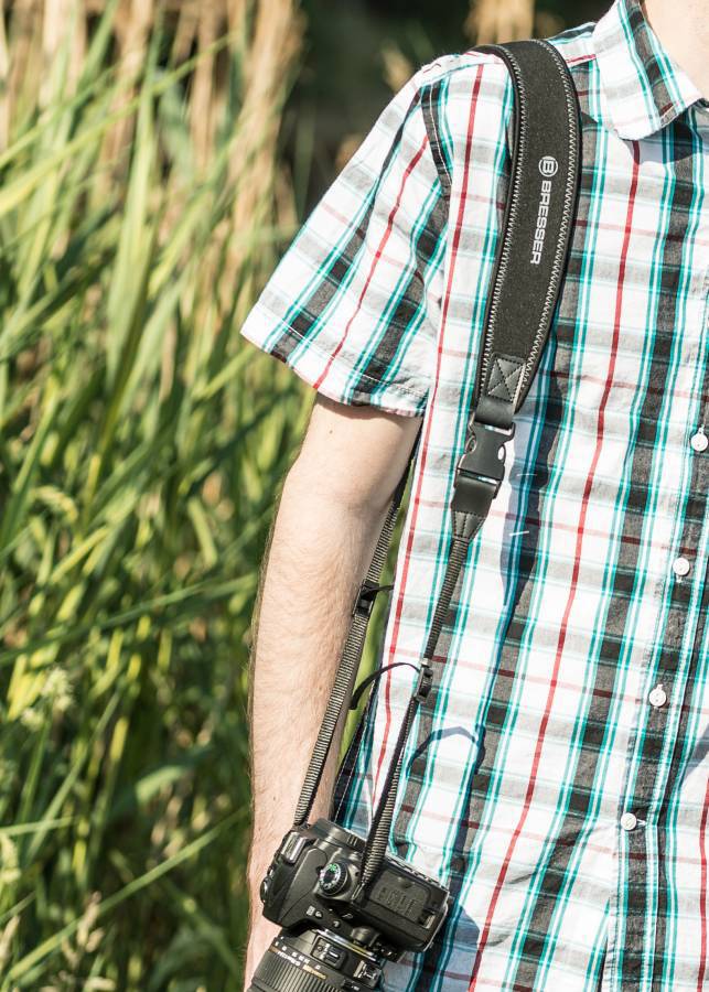 Persoon met geruit overhemd draagt camera met schouderband, buiten, omringd door groene planten.