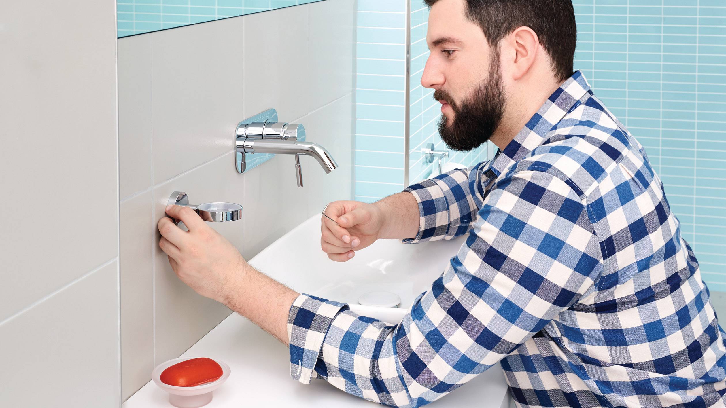 Een man in een geruit shirt repareert een waterkraan in de badkamer. Op de wastafel staat een kom met gereedschappen.