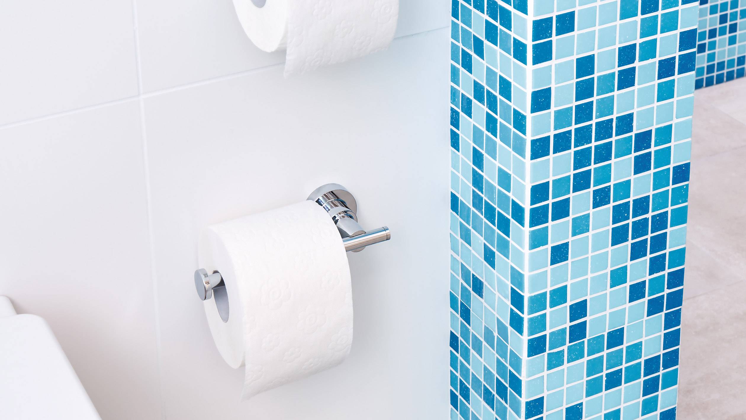 Toiletpapierhouder aan een blauwe betegelde muur. Twee rollen zijn zichtbaar, waarvan er één wordt gebruikt en de andere als reserve dient.