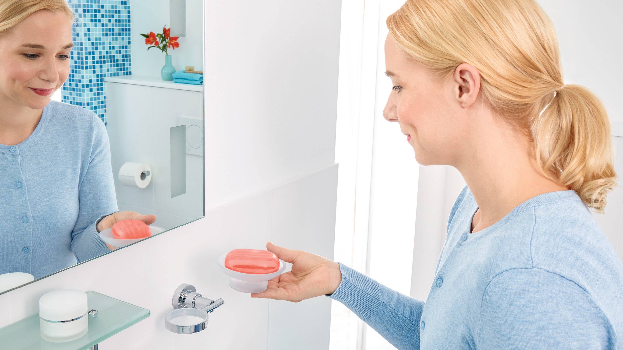 Een vrouw in de badkamer houdt een zeepbakje in haar linkerhand en kijkt naar haar spiegelbeeld terwijl ze haar handen wast.