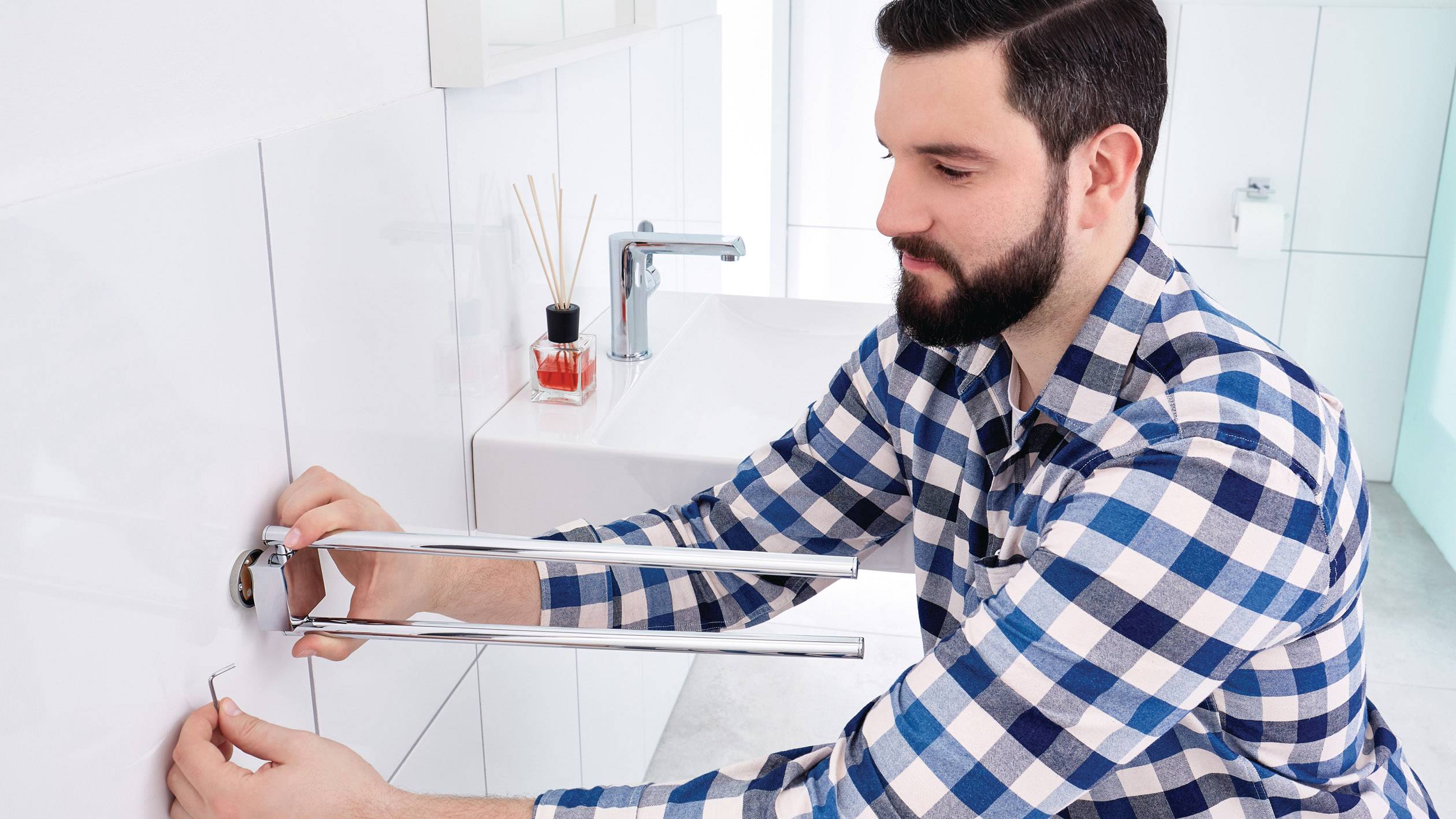 Een man in een geruit overhemd repareert een waterleiding met gereedschap in de badkamer. Op de achtergrond is een wasbak te zien.