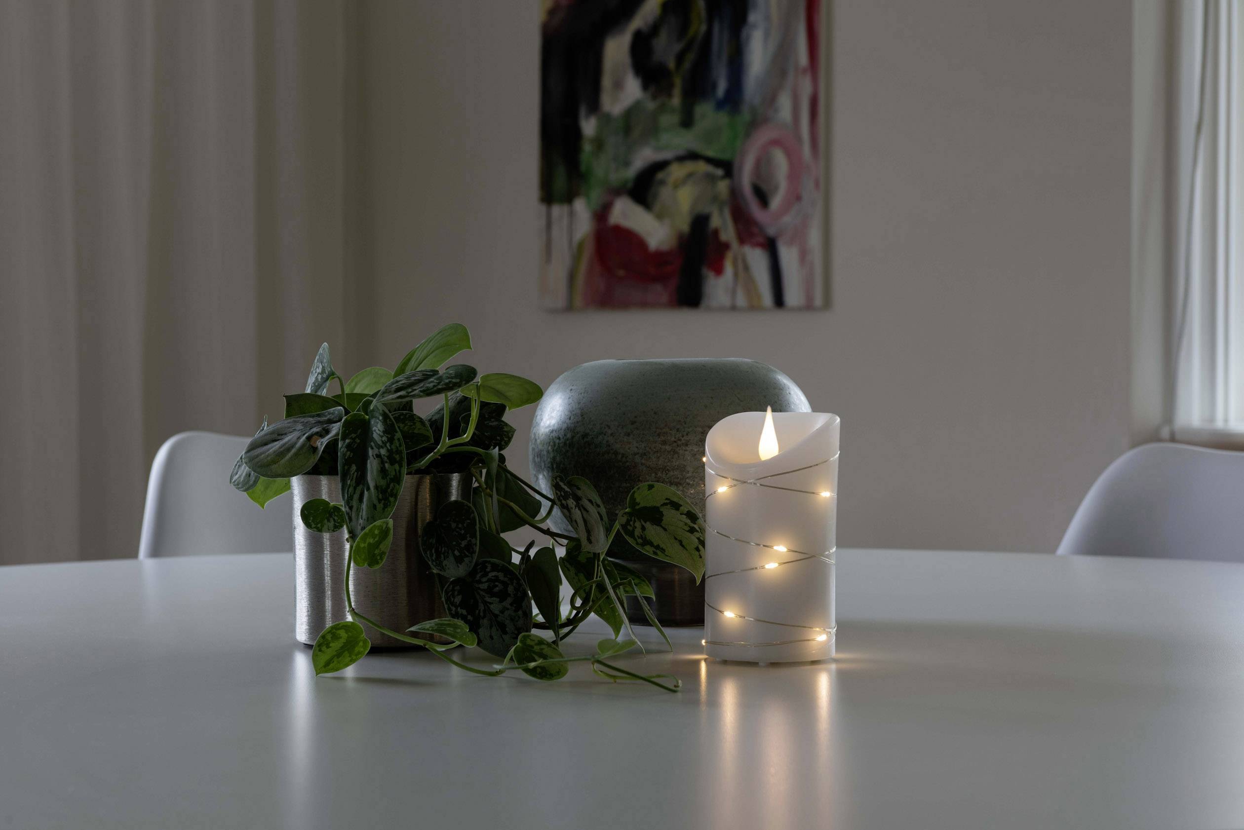Kaars met lichtsnoer op tafel naast planten en kunstwerk aan de muur.