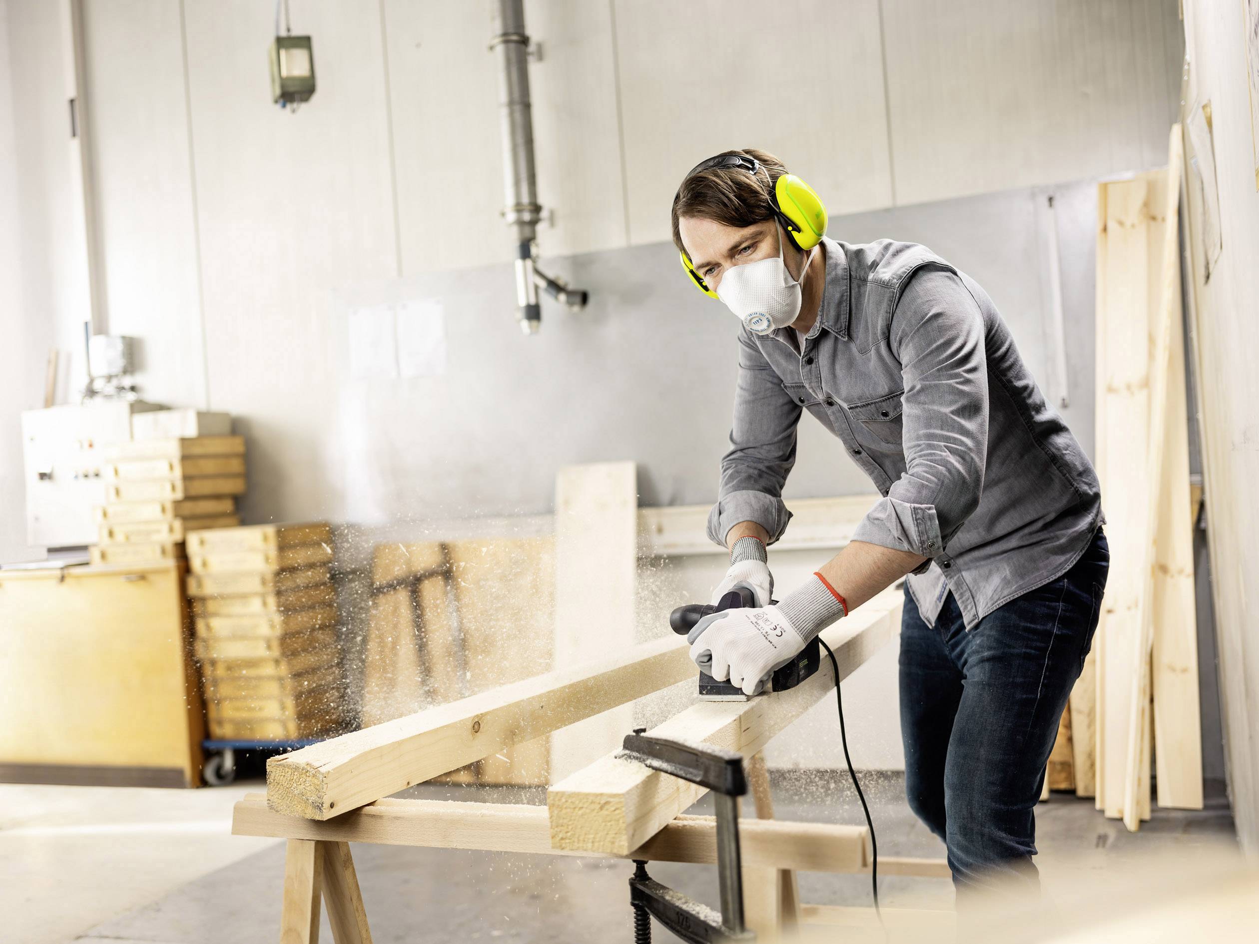 Een man aan het schuren van een houten lat in een werkplaats. Hij draagt een veiligheidsbril, gehoorbescherming en een ademmasker. Houtspaanders vliegen door de lucht.