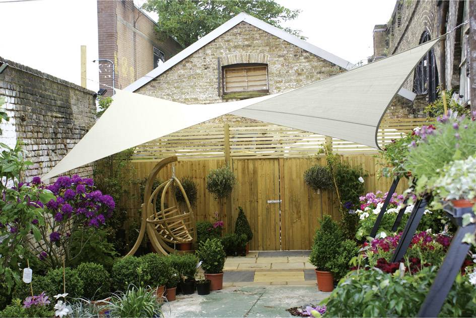 Een kleine achtertuin met bloemen, planten en een houten schommelstoel onder een zonnezeildoek.