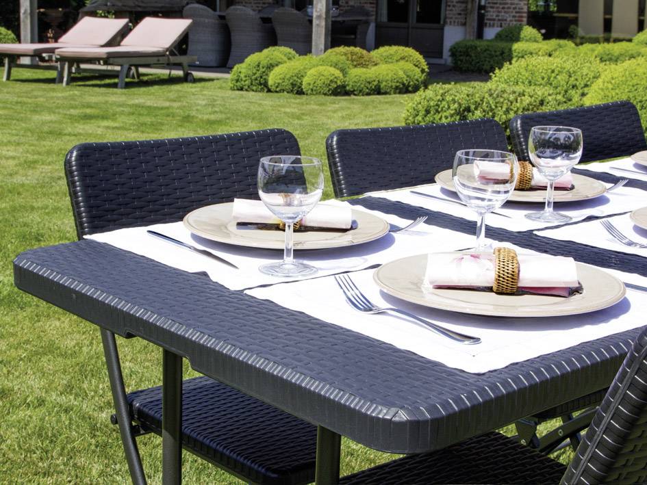 Een gedekte tafel in de tuin met vier glazen, borden en servetten op een zwarte rotan tafel, omgeven door een groen grasveld.