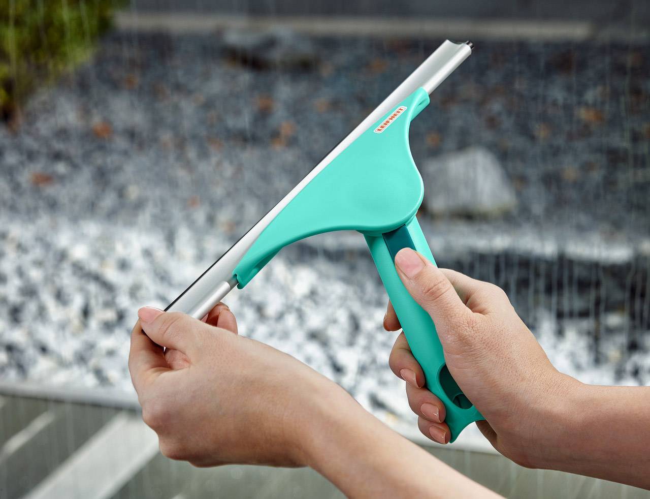 Een persoon houdt een turquoise raamwisser met beide handen boven een grindachtergrond.