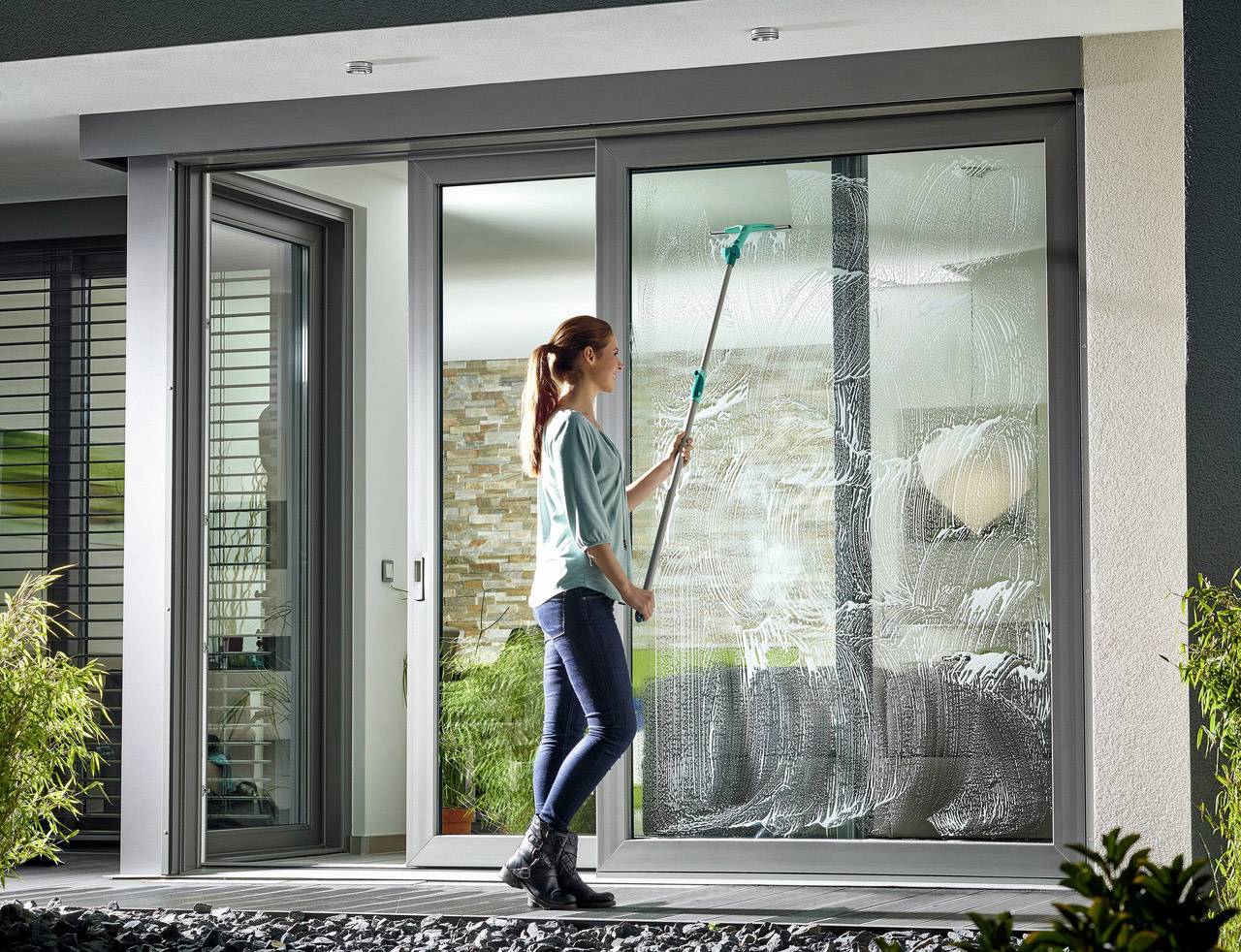 Een persoon reinigt de grote glazen schuifdeur van een modern huis met een stok. Op de achtergrond zijn planten en een stenen muur.