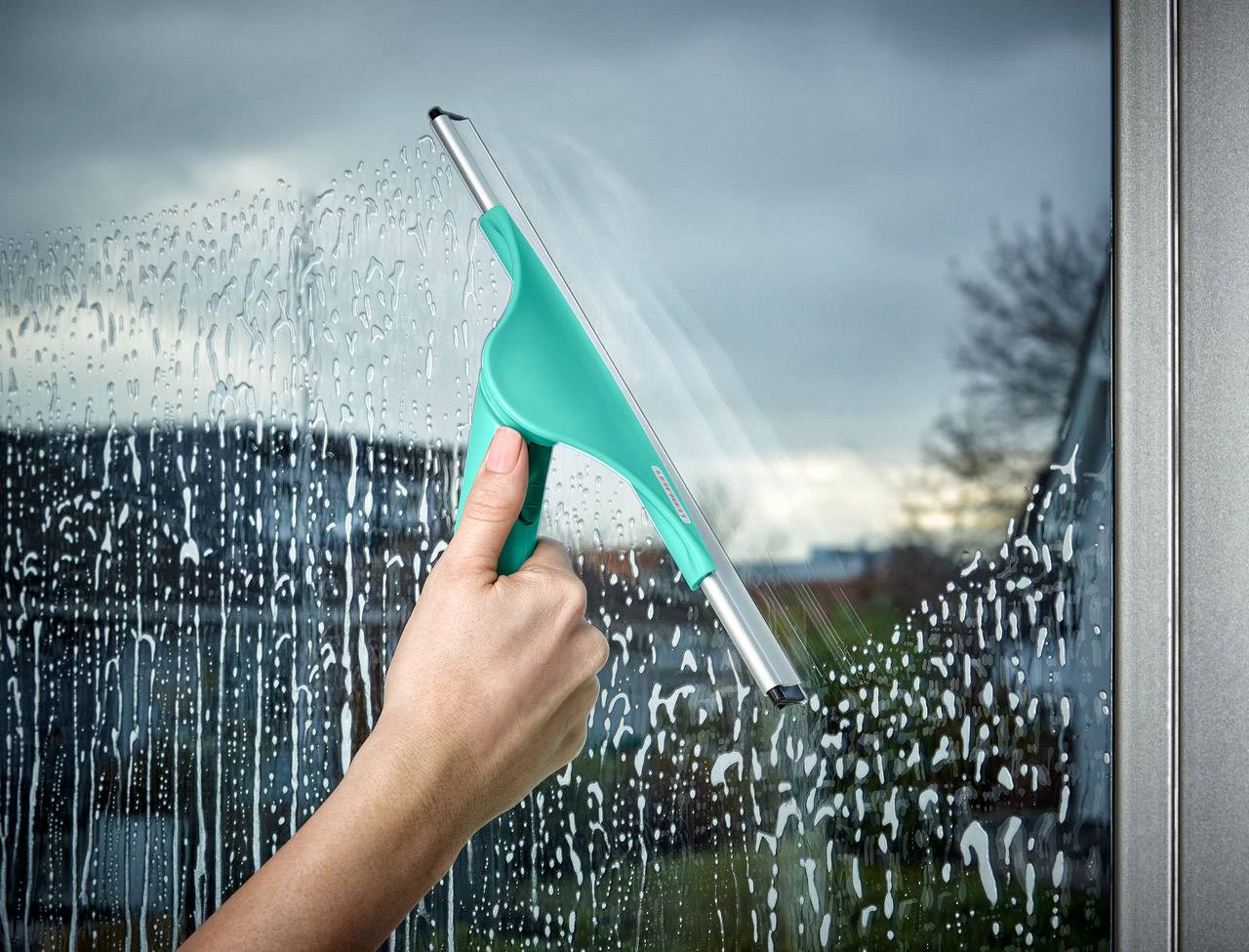 Een persoon reinigt een raam met een groene raamwisser. Zeepwater loopt in strepen over de glasruit.