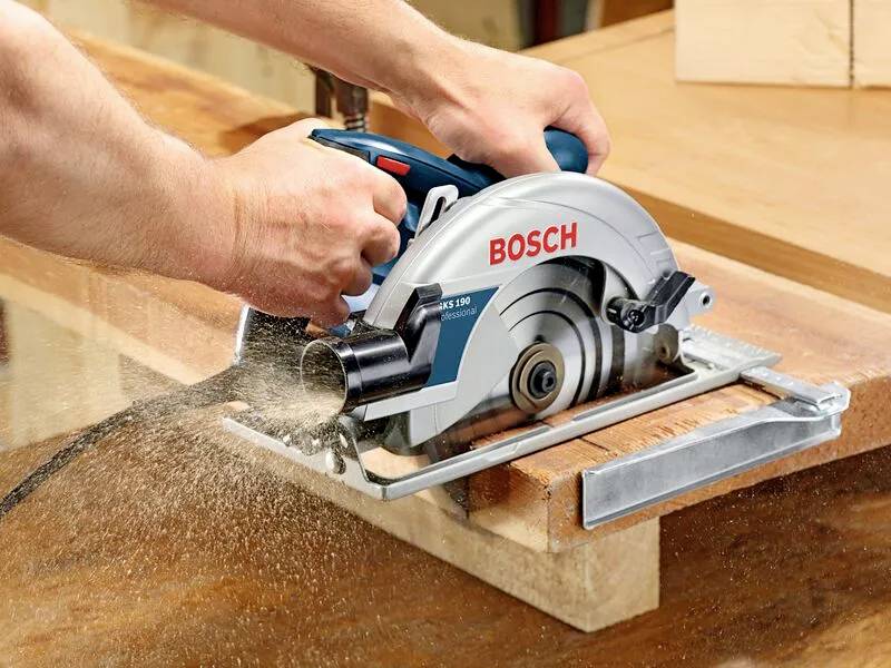Een persoon gebruikt een Bosch cirkelzaag om een houten plank door te zagen. Zaagsel is zichtbaar terwijl het zaagblad door het hout beweegt, waardoor de handeling van het gereedschap wordt geaccentueerd.