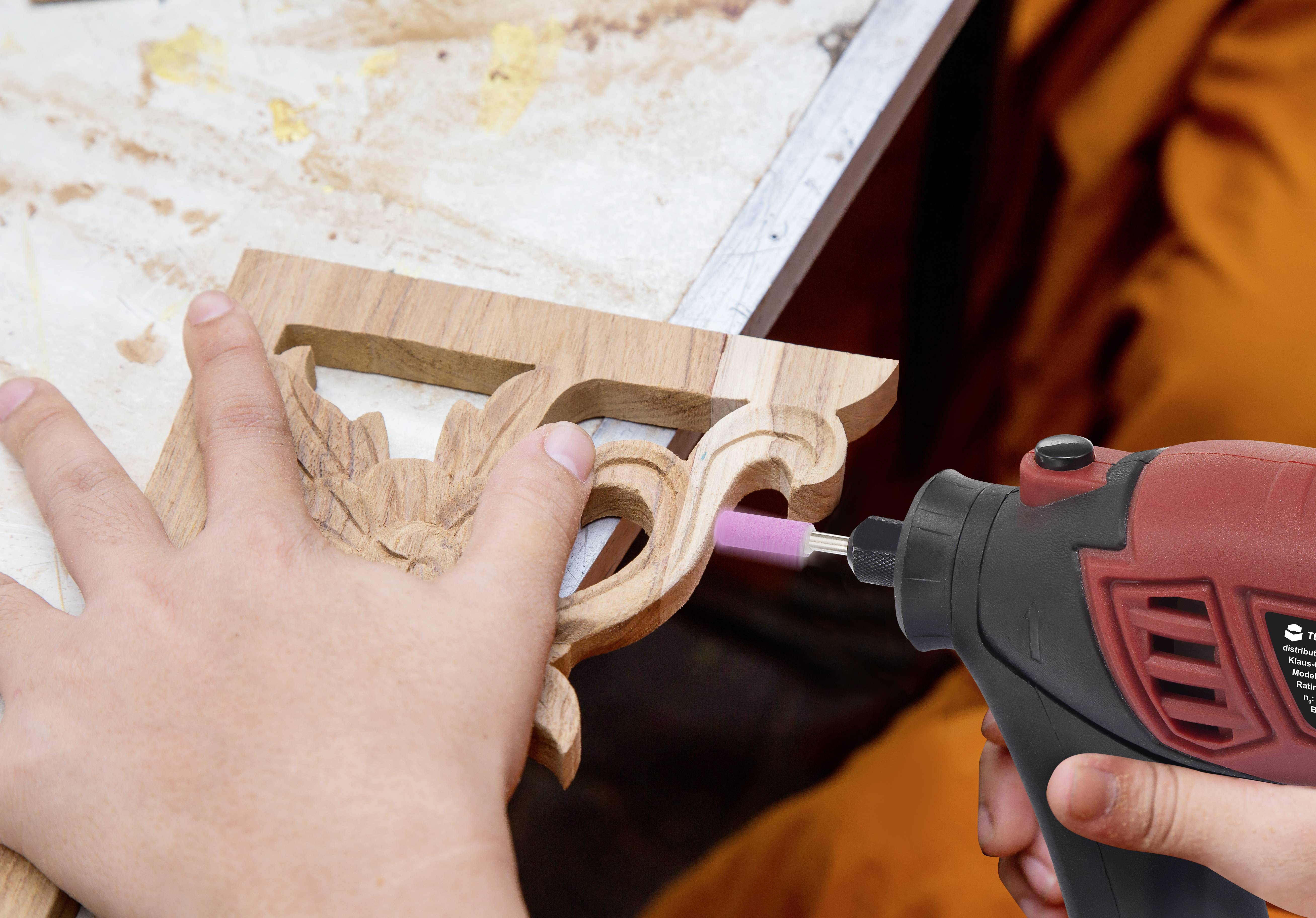 Een persoon bewerkt een houten sculptuur met een rood-zwart slijpgereedschap op een tafel. De handen houden het werkstuk stabiel.
