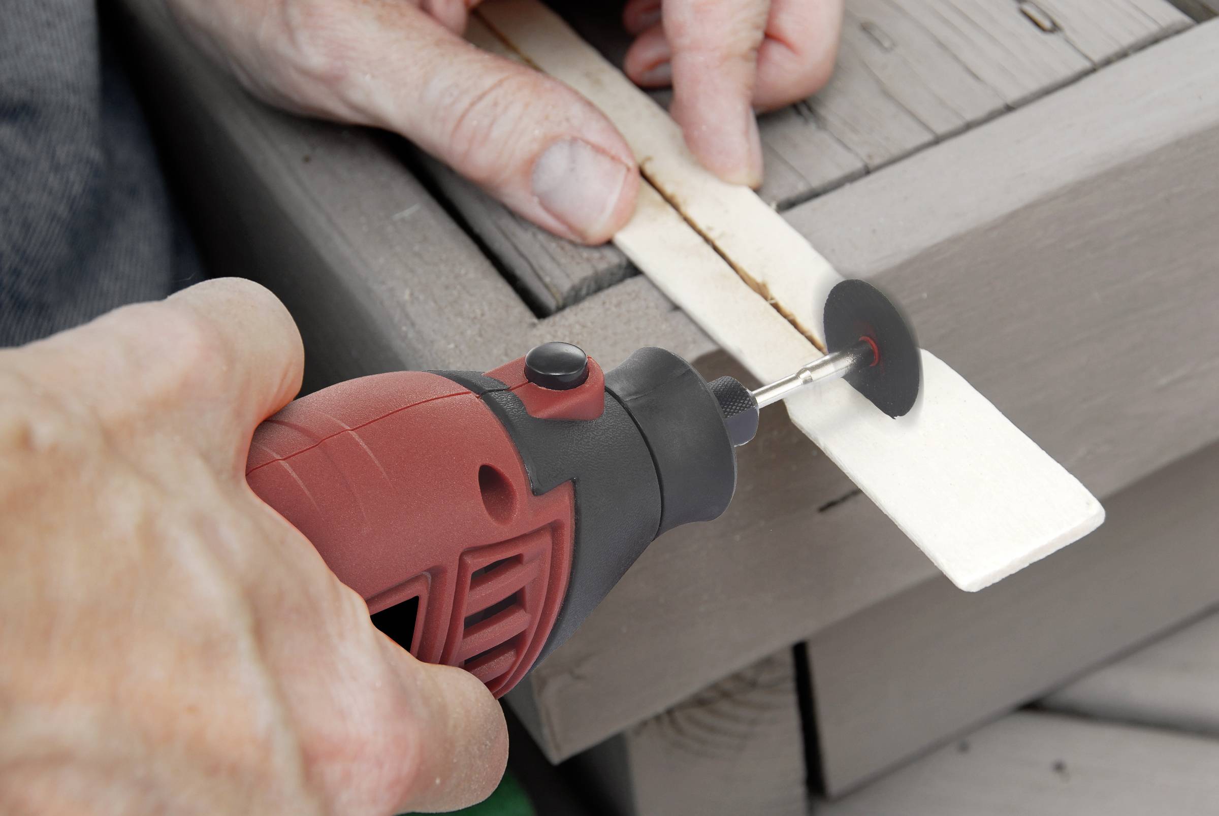 Een persoon snijdt met een rood-zwarte handgereedschap een lange, dunne houten stok op een houten tafel.