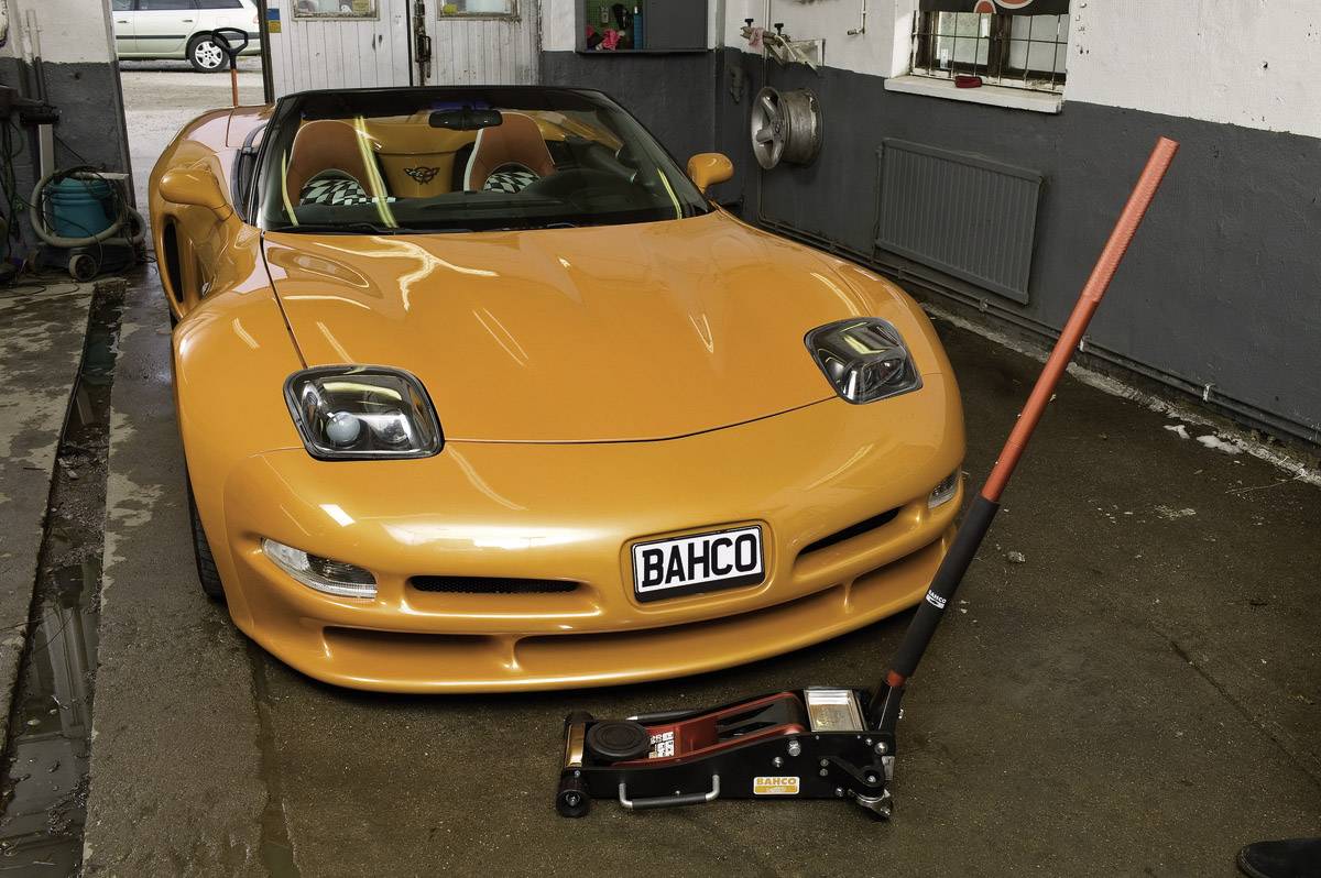 Oranje sportwagen in een werkplaats met een krik ervoor. Nummerplaat toont 'BAHCO'.